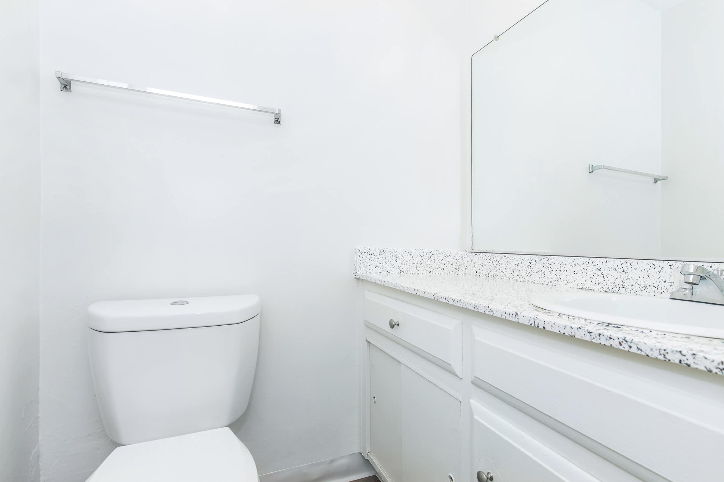 A clean and modern bathroom featuring a white toilet, a sink with a granite countertop, and a large mirror. The walls are painted white, and there is a towel bar next to the mirror, creating a bright and spacious feel.