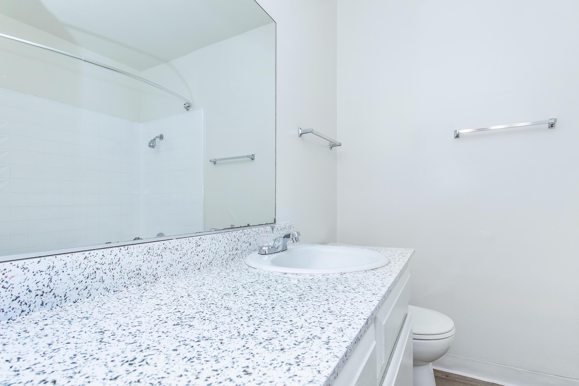 A clean and modern bathroom featuring a white countertop with a speckled pattern, a round sink with a chrome faucet, a large mirror above the sink, and a toilet in the background. The walls are painted white, and there are towel bars installed on either side of the mirror. Natural light brightens the space.
