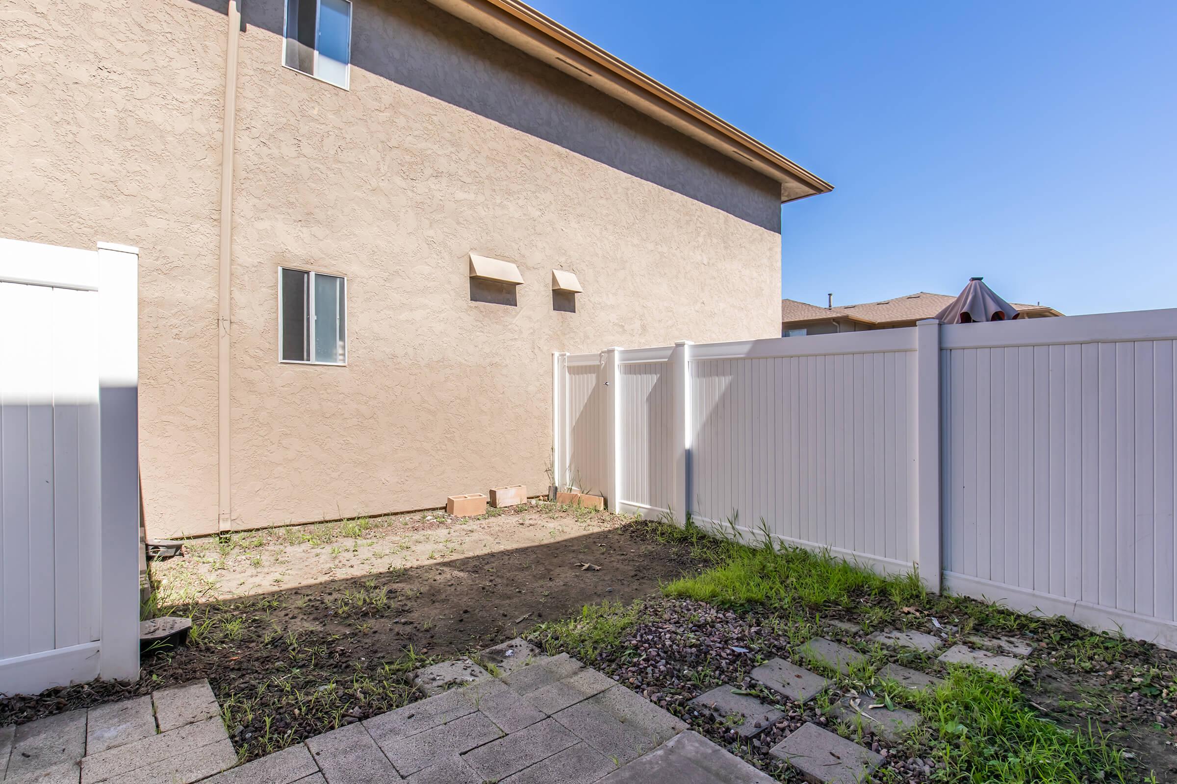 A view of a bare backyard space enclosed by a white vinyl fence, featuring a light-colored stucco wall of a house in the background. There are patchy areas of grass and a few concrete pavers on the ground, with clear blue skies overhead. The area appears to be well-maintained but empty.