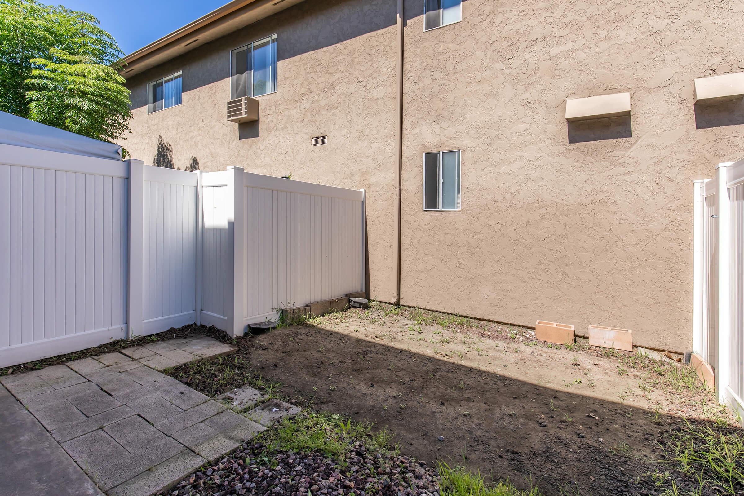 A small, empty backyard area featuring a light-colored wall, a white fence, and a patch of dirt with some sparse grass. There are a few small bricks on the ground and a window visible on the adjacent building. The scene is well-lit with clear blue skies in the background.