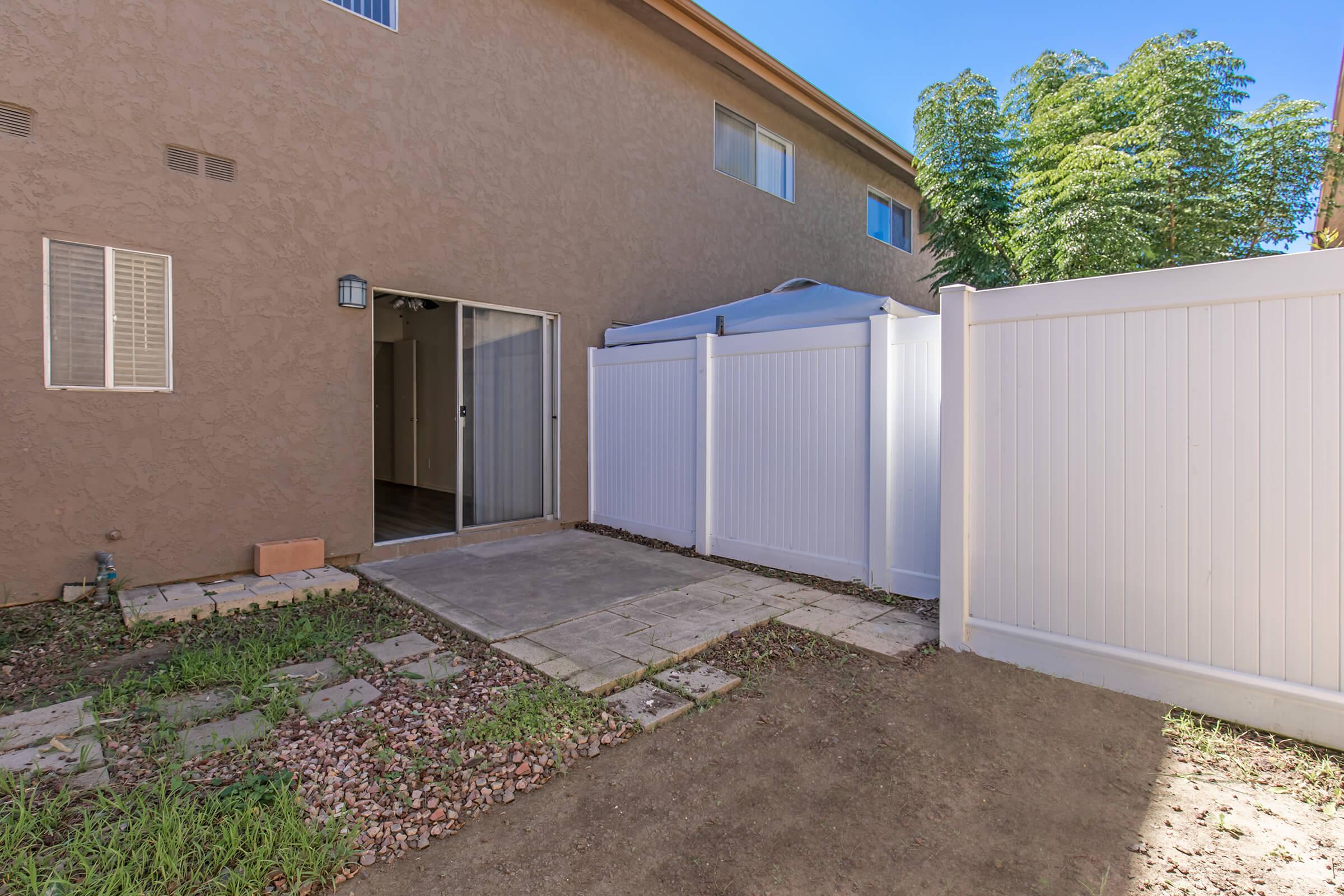 A backyard patio area featuring a small stone pathway, surrounded by gravel and grass. A white fence encloses the space, with a sliding glass door leading into the house. The area is well-lit, with greenery in the background and a clear blue sky above.