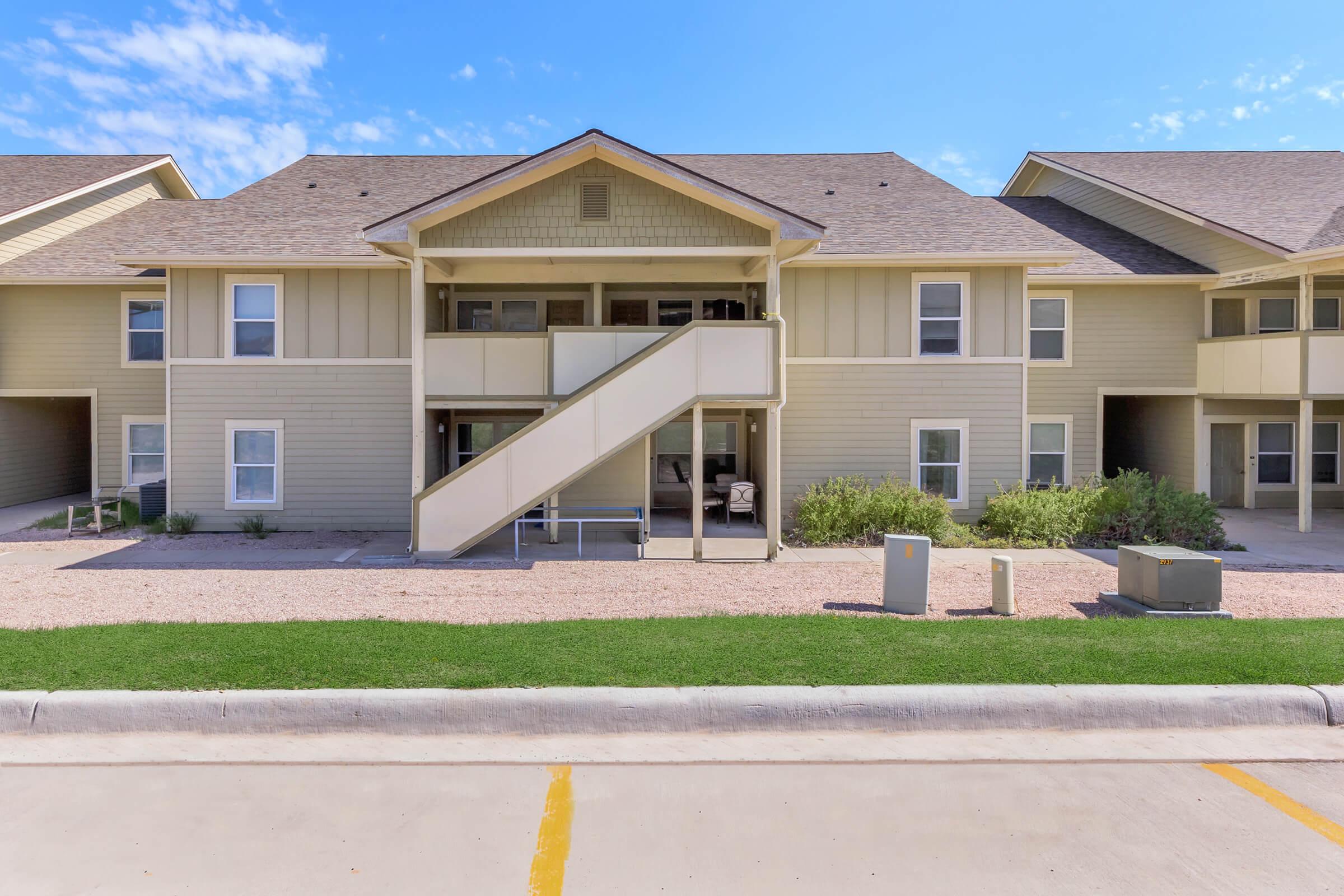 Two-story residential building with beige siding, featuring a central staircase leading to upper units. The ground level has a patio area, and the surrounding landscape includes grass and gravel. The sky is clear with a few clouds.