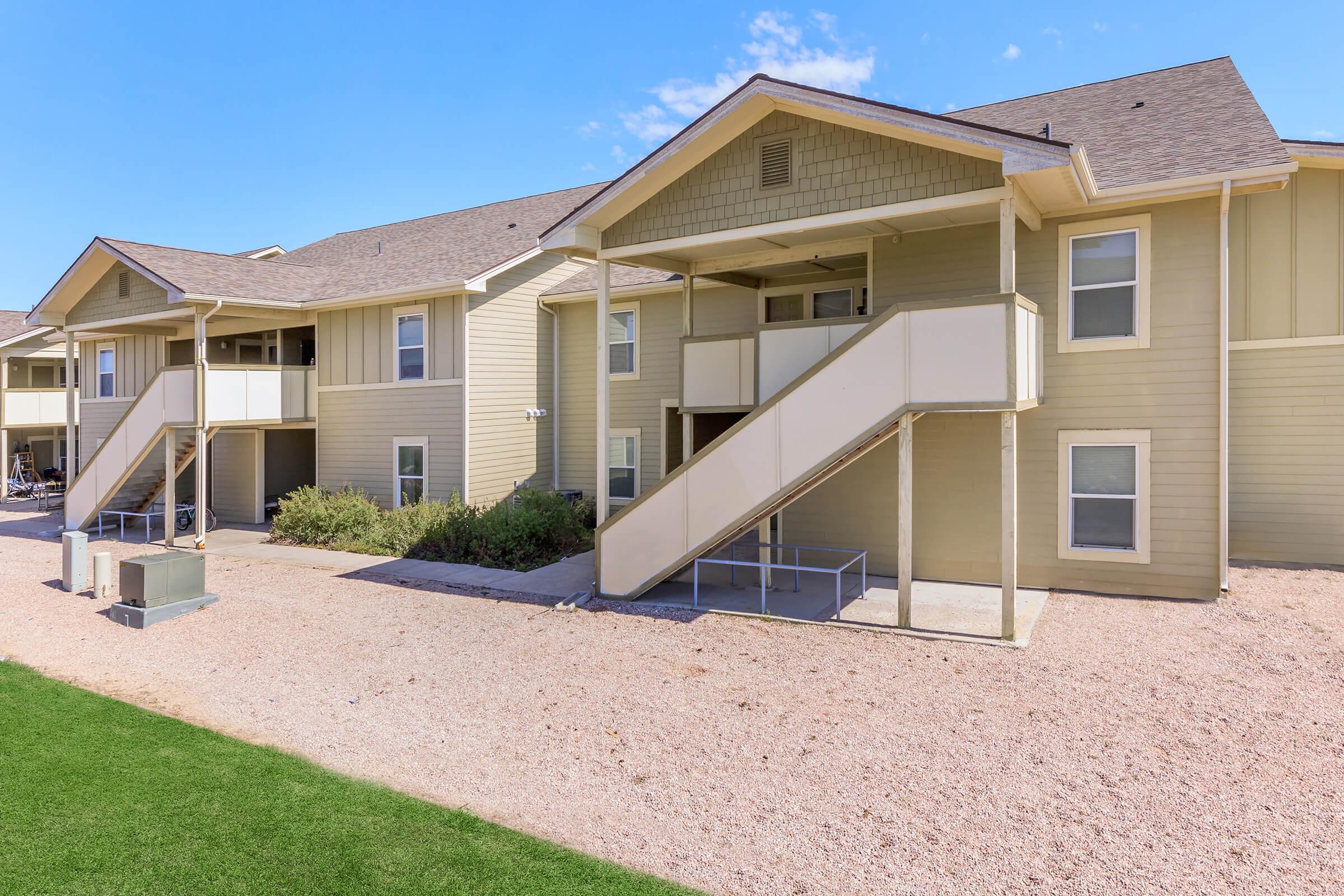 Two-story residential building with beige siding and sloped roofs. Balconies with stairs lead up to apartments. A gravel pathway runs in front, with green grass on one side and utility equipment visible. Clear blue sky in the background.