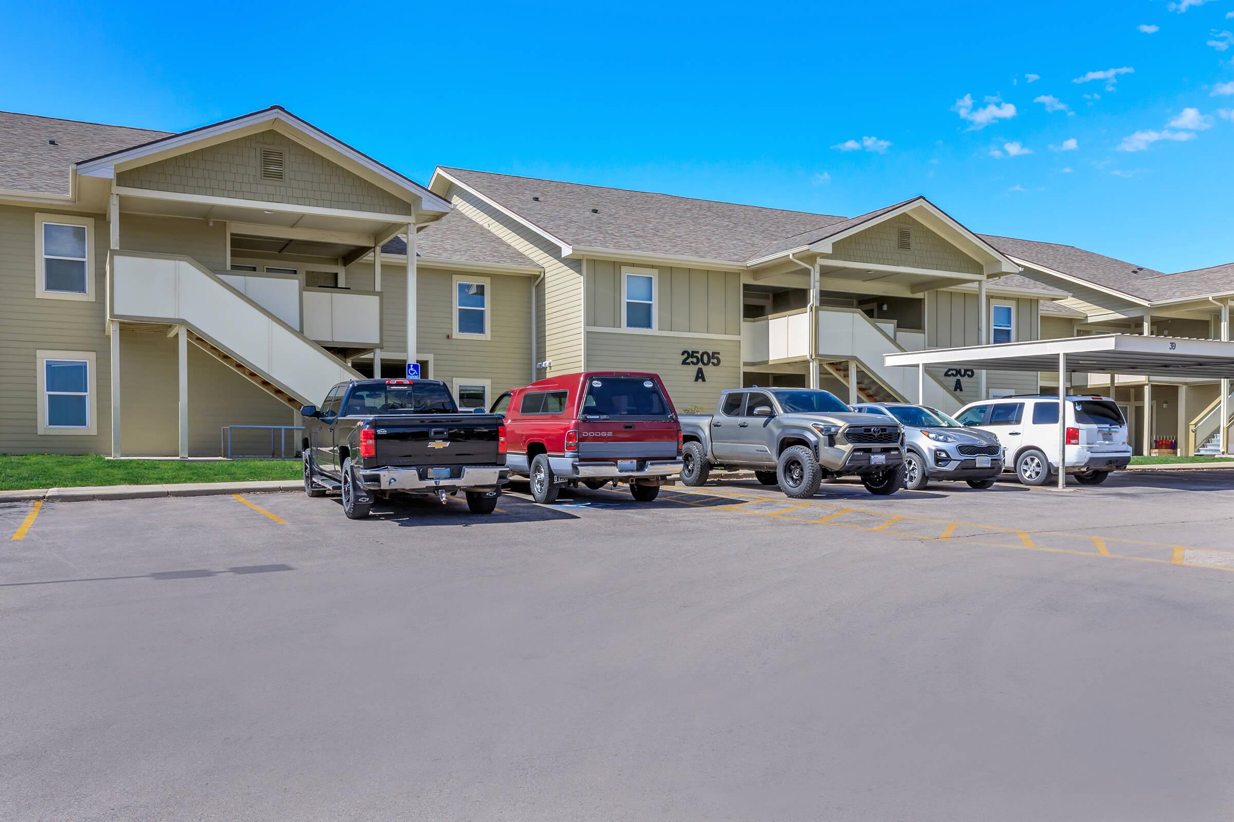 Exterior view of a two-story apartment building with a light yellow facade. Several vehicles are parked in the foreground, including a black truck, a red truck, and an SUV. Clear blue sky and well-maintained grass surround the building, which features outdoor staircases leading to the second floor.