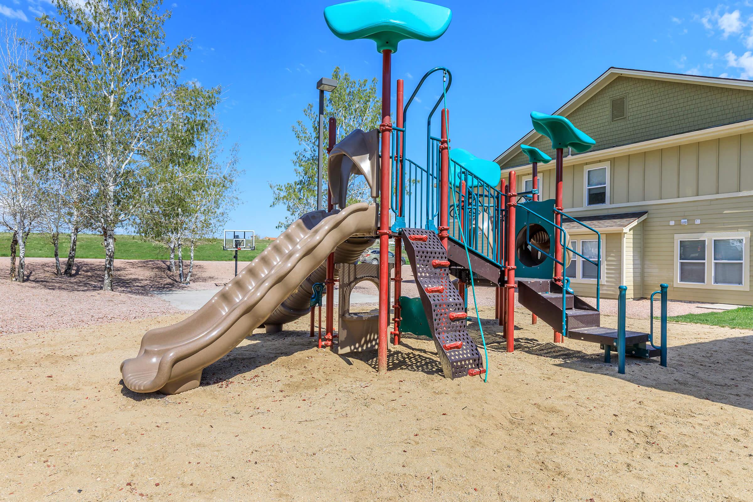 A colorful playground featuring a tan slide, climbing structures, and various play features, set on sandy ground with trees in the background. A building is visible nearby, creating a scenic outdoor play area.