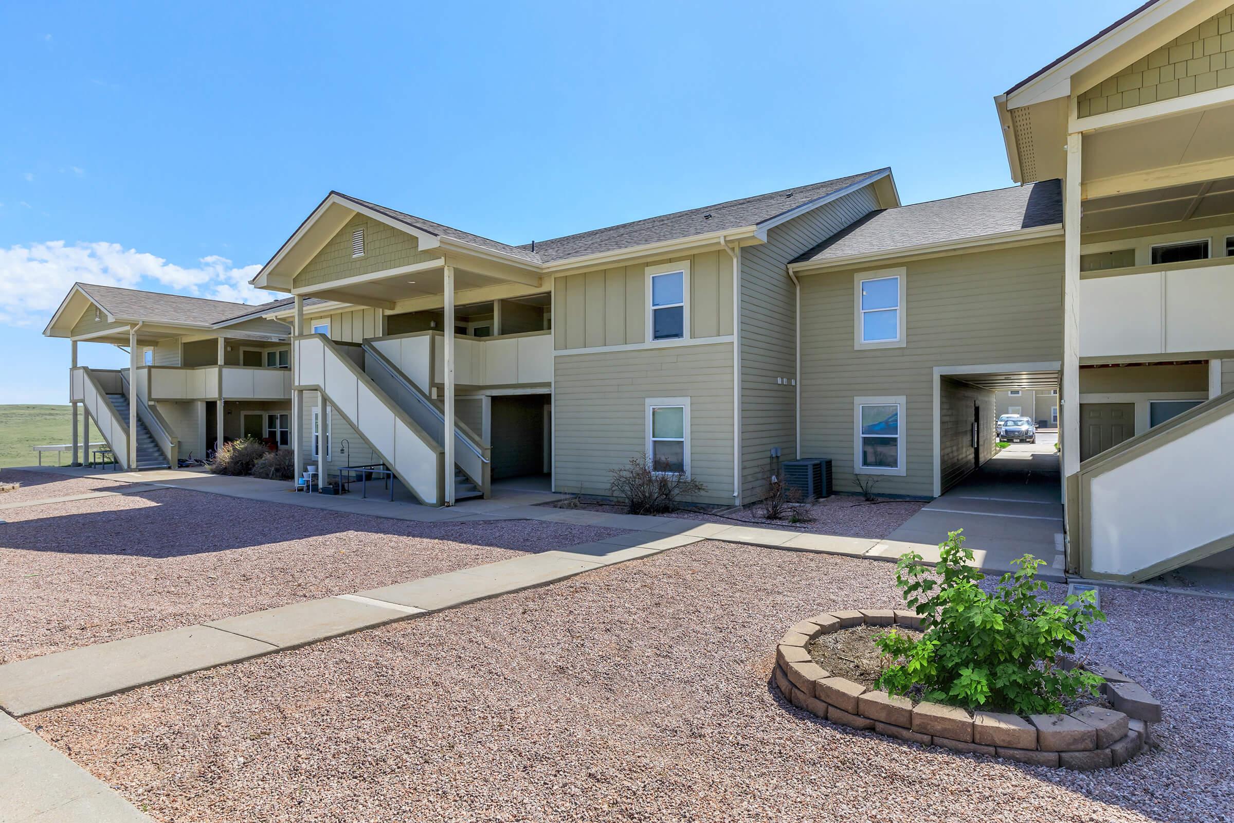 Two-story apartment building with multiple units featuring porches and staircases. The walkway is paved, bordered by small rocks and a circular flower bed with a green plant. Clear blue sky visible in the background, indicating a sunny day.