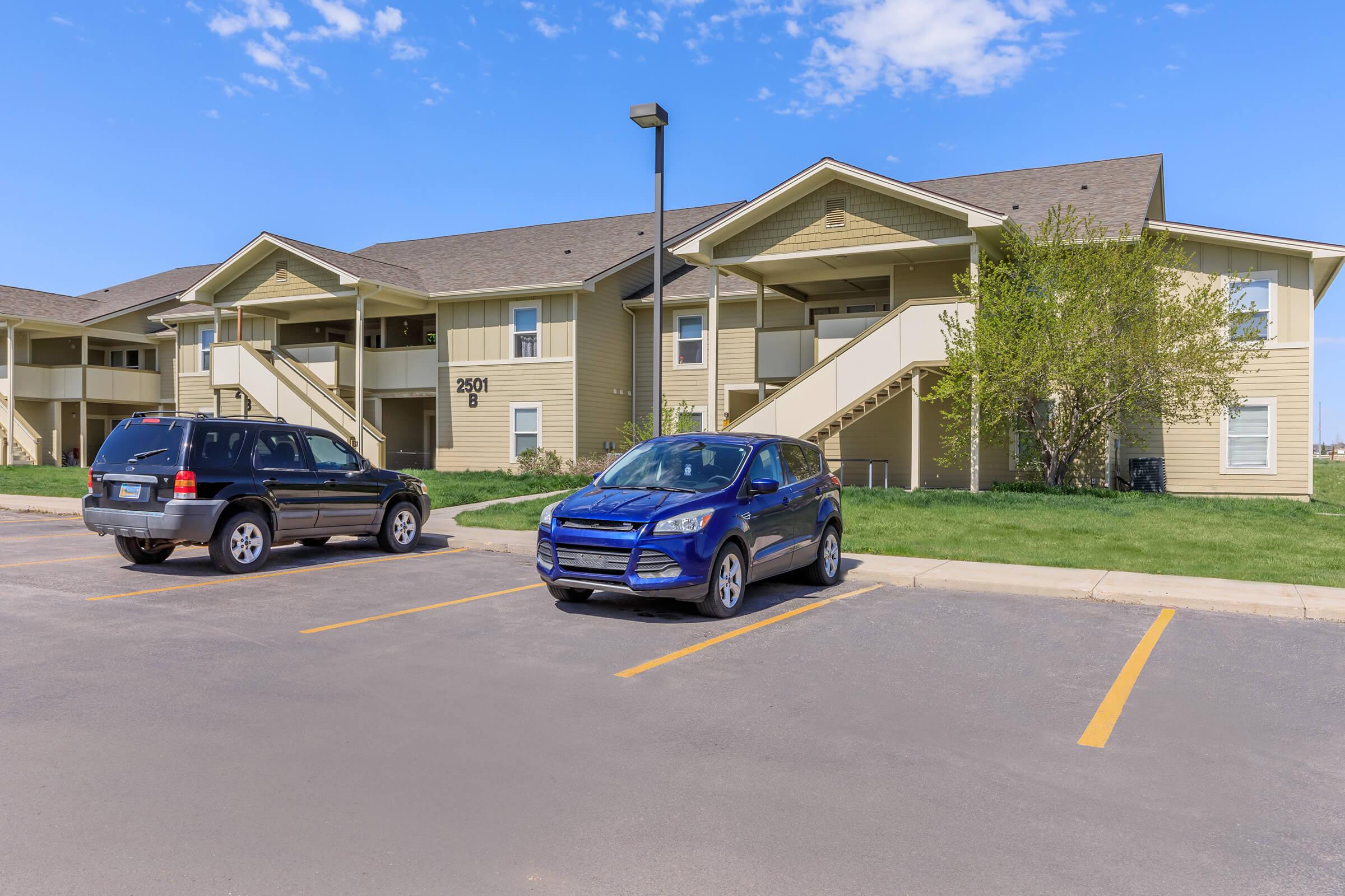Two parked cars, a black SUV and a blue compact vehicle, sit in front of a two-story apartment building with a light-colored exterior and green lawn. Stairs lead to the second level of the building. The sky above is clear with a few clouds, suggesting a pleasant day.