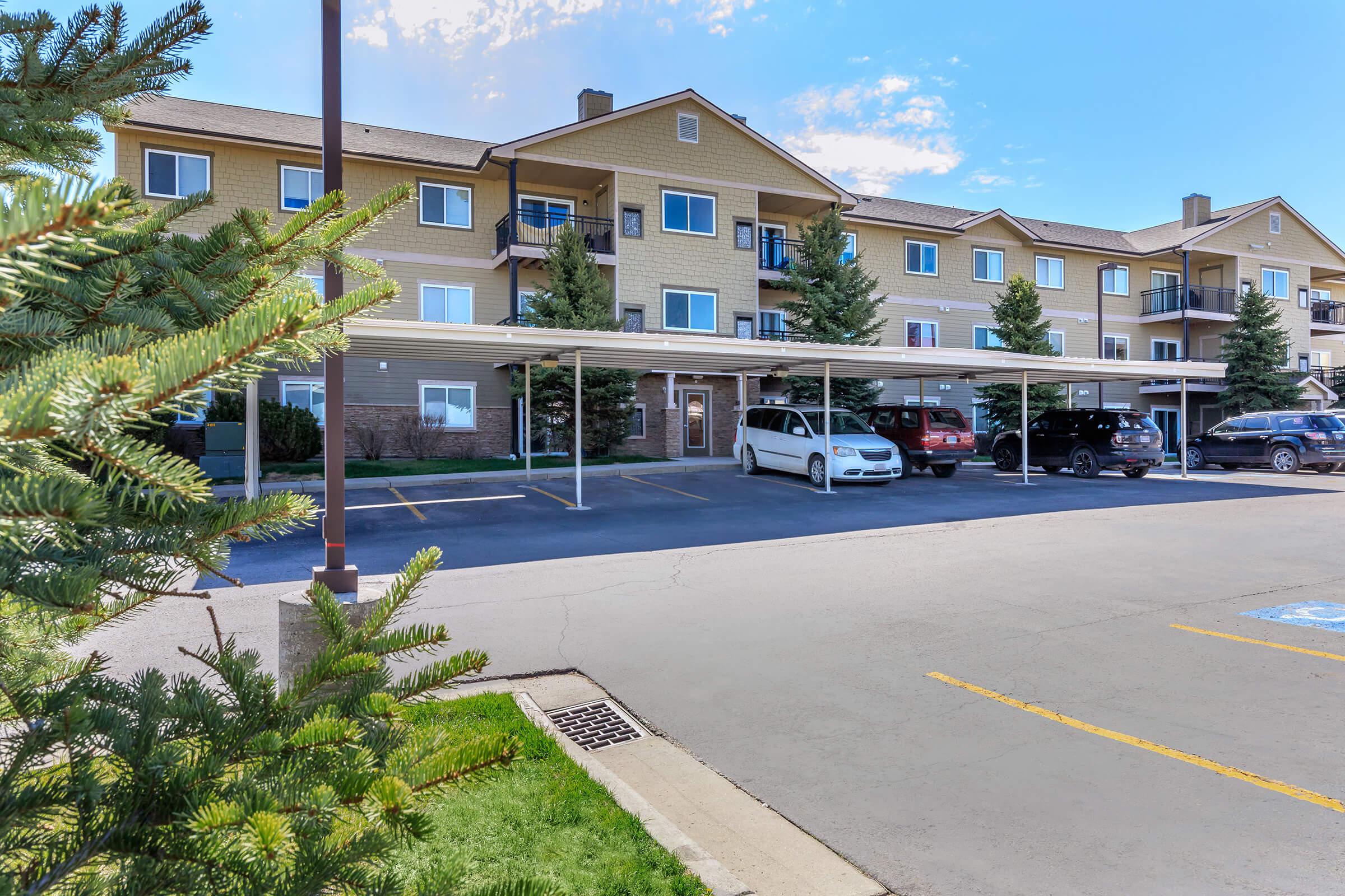 Residential building with multiple apartments visible, featuring covered parking spaces in the foreground. Green trees are present in the image, and a clear blue sky is seen overhead. The area appears well-maintained, with a paved driveway and parking lot.