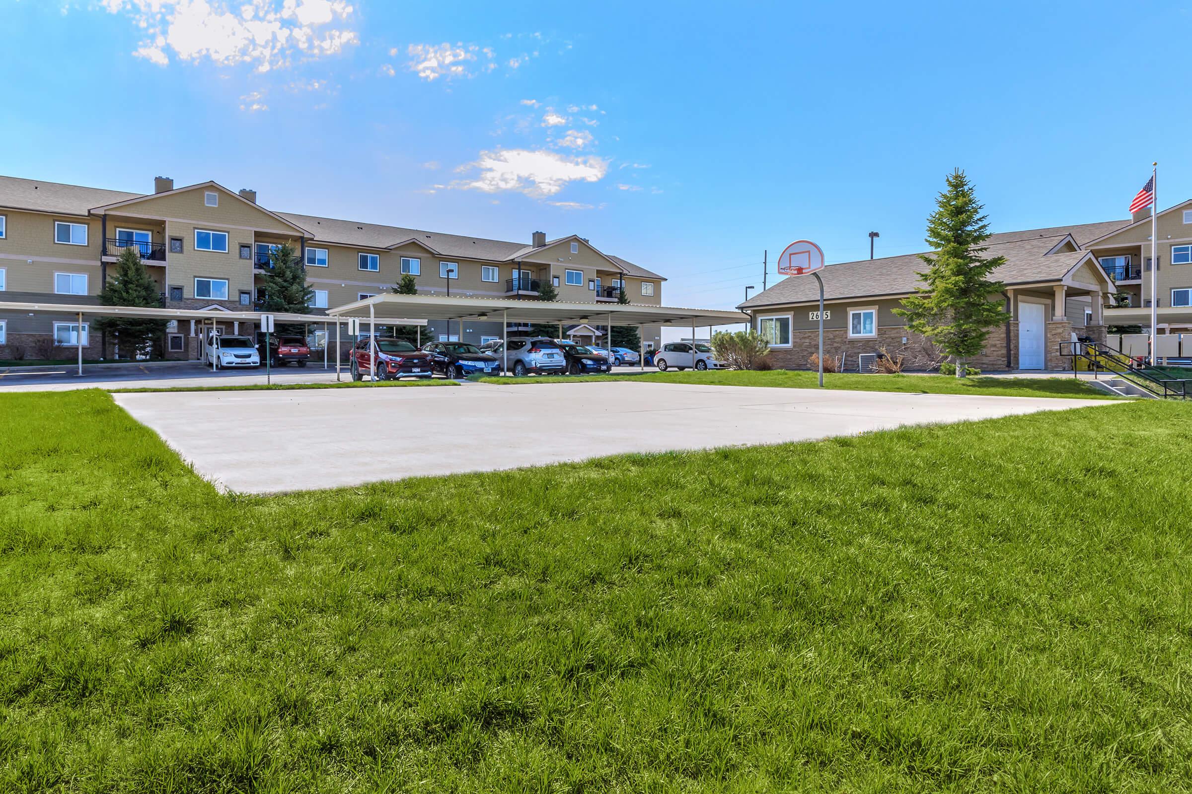 A well-maintained grassy area features a concrete basketball court in the foreground. In the background, there are two multi-story residential buildings with covered parking. A basketball hoop is visible near the court, and an American flag is displayed on one of the buildings under a clear blue sky.