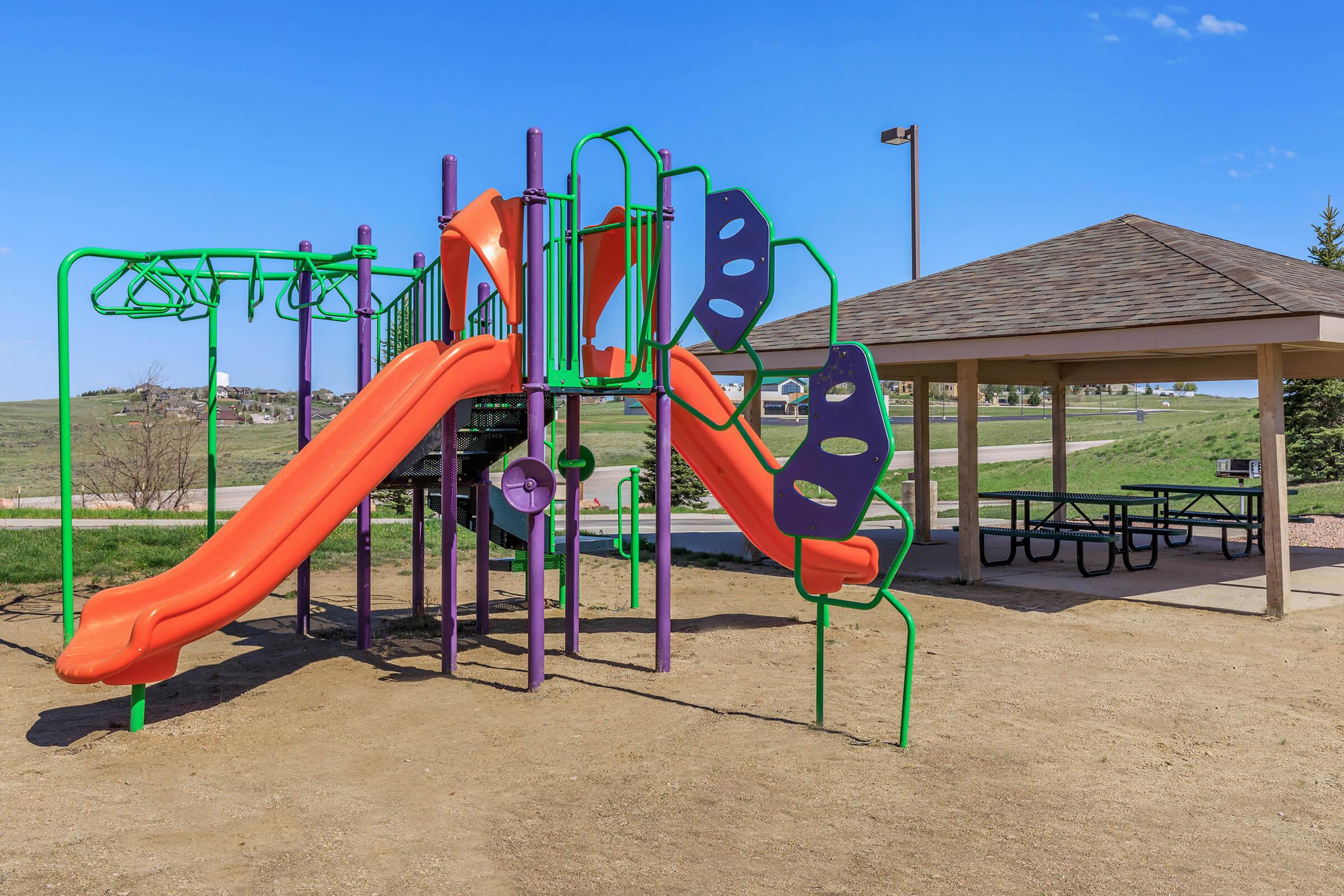 A colorful playground structure featuring orange slides and green climbing elements, set on sandy ground. Nearby, a shaded picnic area with a wooden gazebo and picnic tables can be seen against a backdrop of open grassland and a clear blue sky.