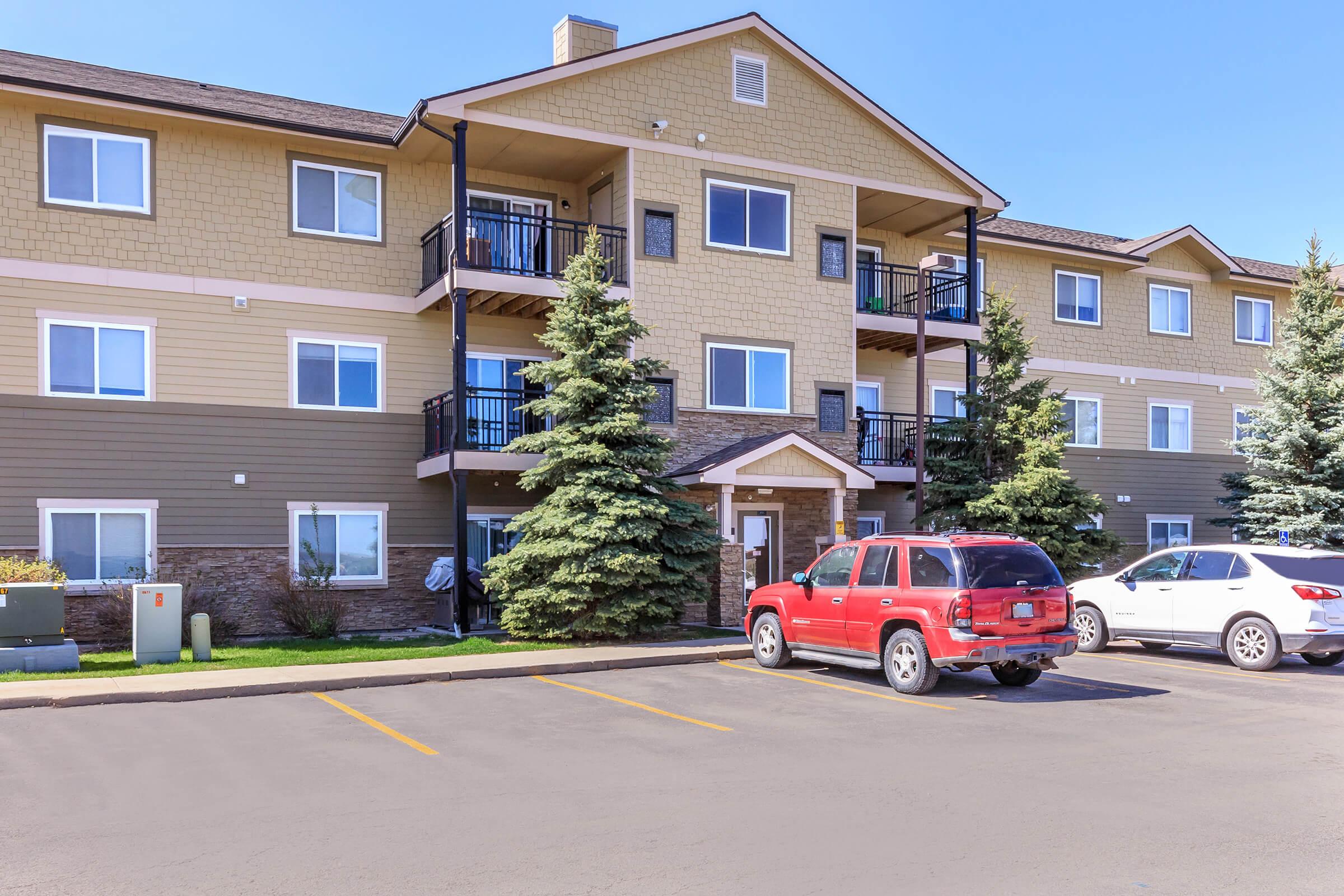 Multi-story apartment building with a beige exterior, featuring balconies and large windows. Two evergreen trees are planted in front. A red SUV is parked in the lot, along with a white vehicle. The sky is clear and sunny, indicating a bright day.