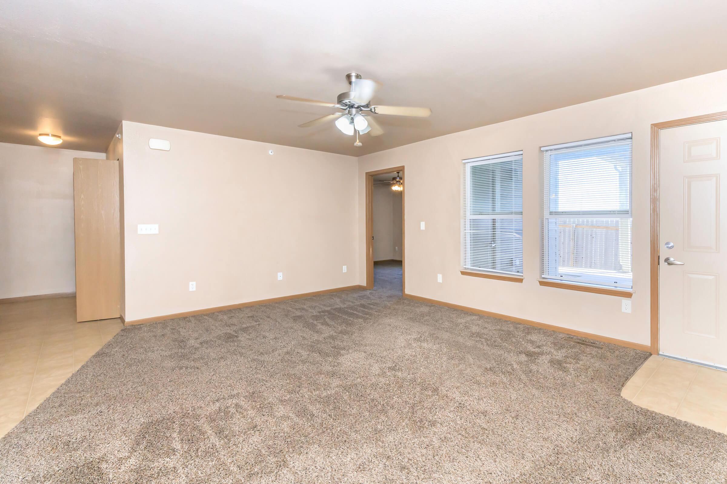 Spacious living room featuring light-colored walls and a ceiling fan. The floor is covered in plush beige carpet, with windows allowing natural light. A doorway leads to another room, and a closed door is visible, suggesting additional space. Overall, the room has a cozy and inviting atmosphere.