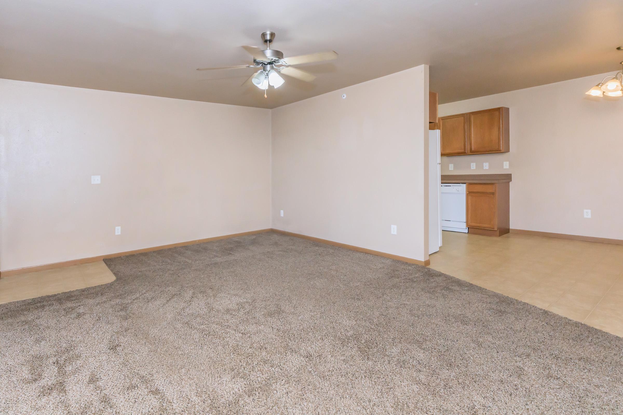 Spacious, light-filled living area featuring a ceiling fan, soft beige walls, and plush carpet. An open layout leads to a kitchen with wooden cabinets and white appliances, enhancing the inviting atmosphere. Ideal for comfortable living and entertaining.