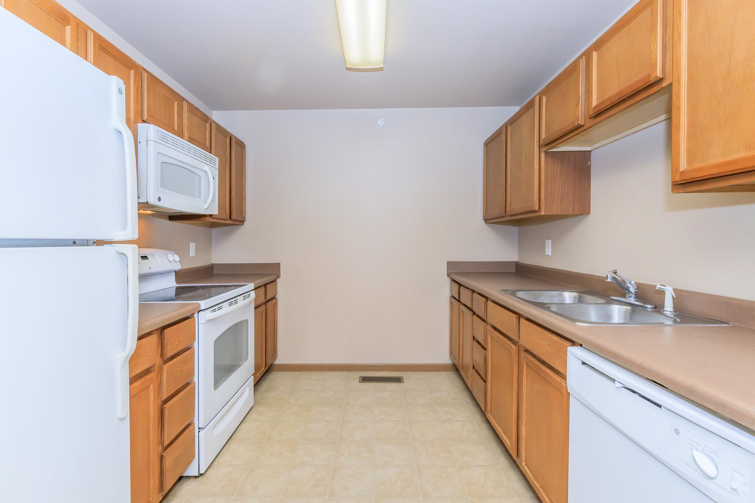 A clean, modern kitchen featuring wooden cabinetry, white appliances, and a neutral wall color. The layout includes a refrigerator, oven, microwave, and dishwasher, along with double sinks. The floor is tiled, creating a spacious and inviting atmosphere.