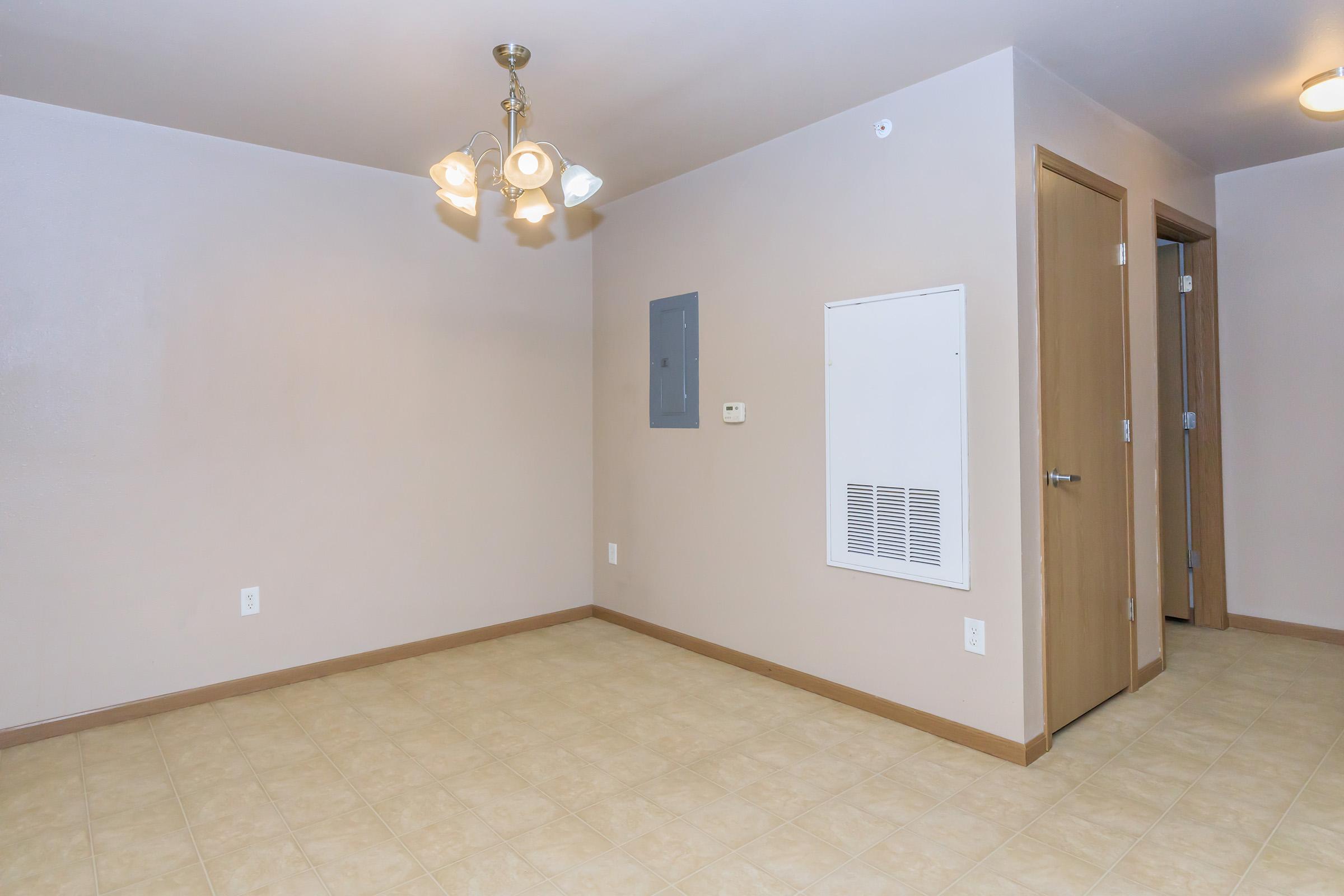 Empty room featuring beige walls and light-colored tile flooring. A ceiling light fixture with multiple bulbs hangs from the ceiling. To the right, there is a closed door and an electrical panel mounted on the wall. The overall space appears well-lit and ready for furnishing.
