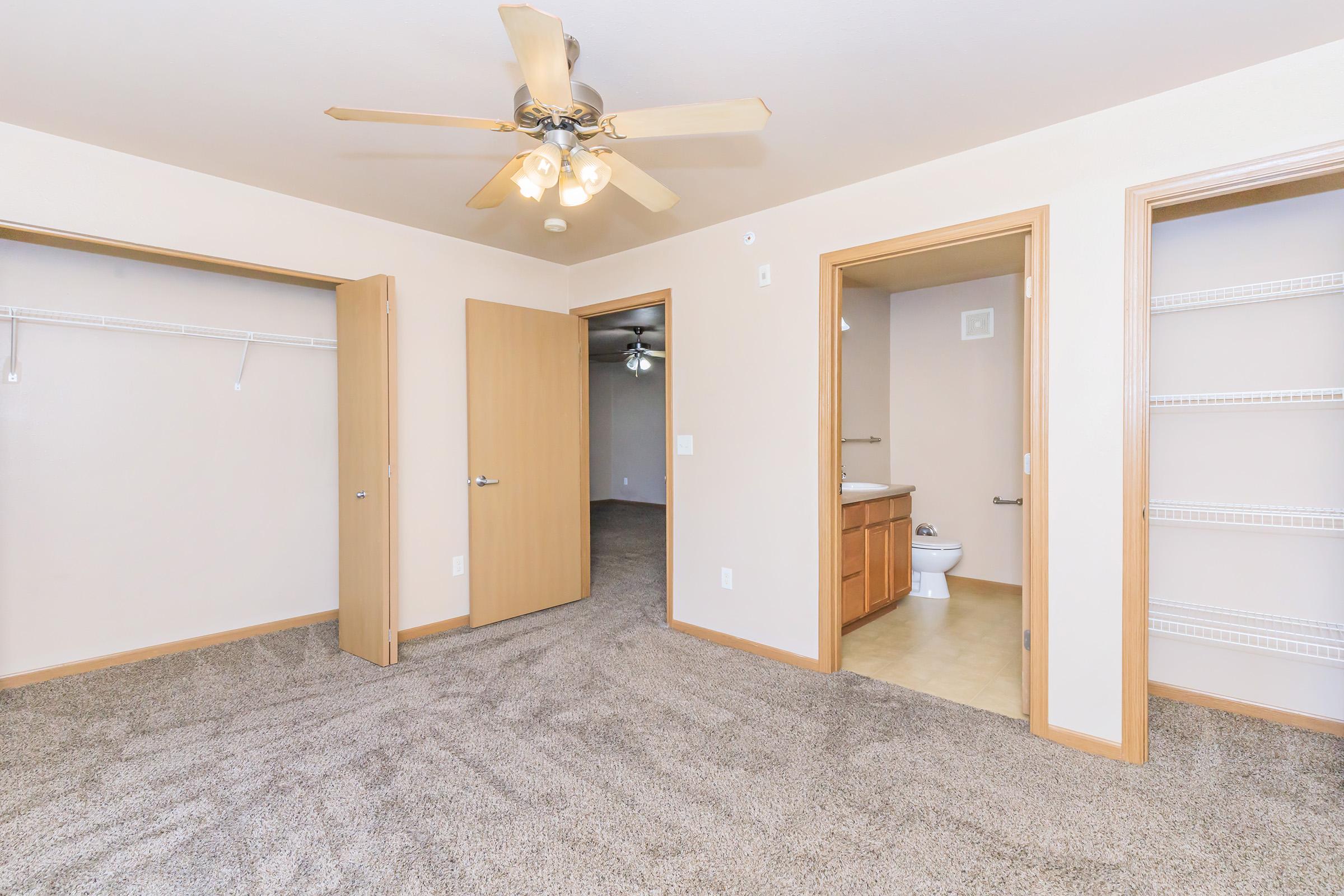 Interior view of a spacious room featuring a ceiling fan, beige walls, and soft carpet flooring. There are two open doorways leading to other areas, a bathroom visible in the background, and multiple closet spaces with shelving. Natural light fills the space, creating a welcoming atmosphere.