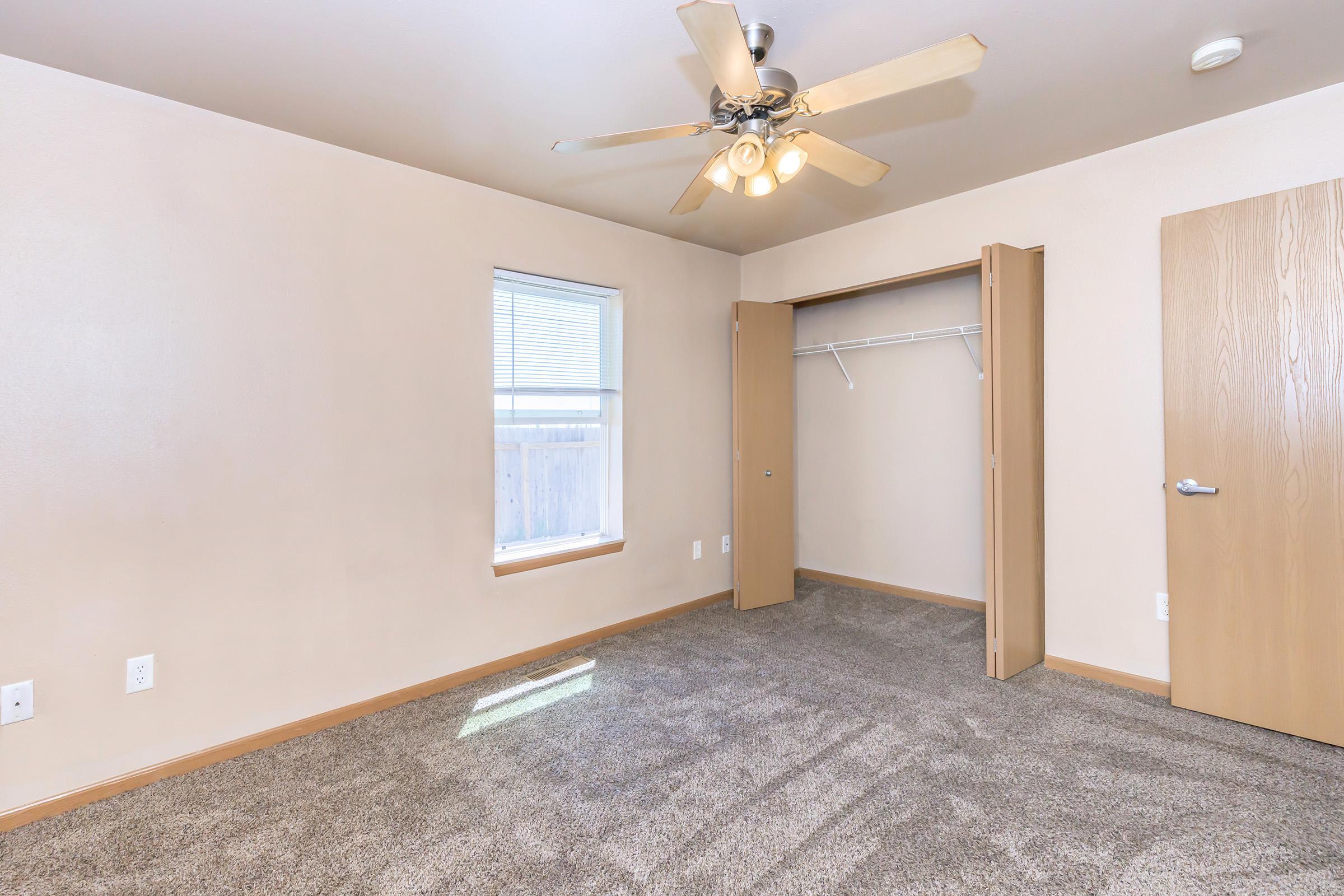 A well-lit empty bedroom featuring beige walls, a ceiling fan with light, and a carpeted floor. One wall has a window with blinds, and there is an open closet with a white rod for hanging clothes. The doors to the closet and room are light wood. The space appears clean and ready for decoration or furniture.