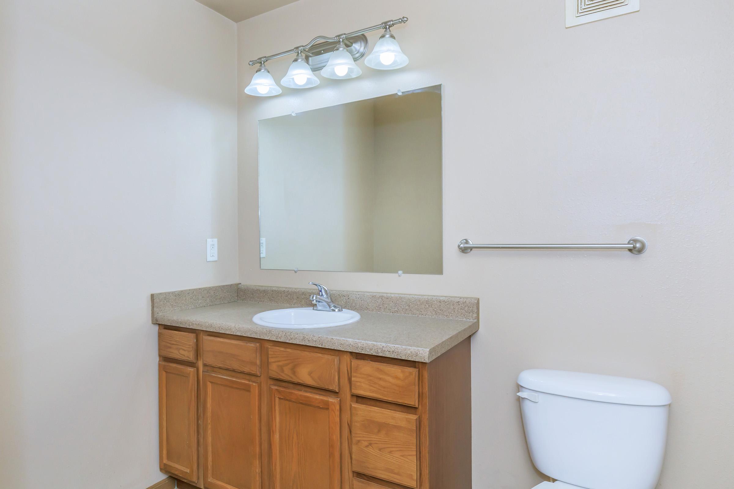 A clean and simple bathroom featuring a wooden vanity with a sink, a large mirror above, a light fixture with four bulbs, and a white toilet. The walls are painted a neutral color, and a towel bar is installed next to the mirror. The overall layout is spacious and well-lit.
