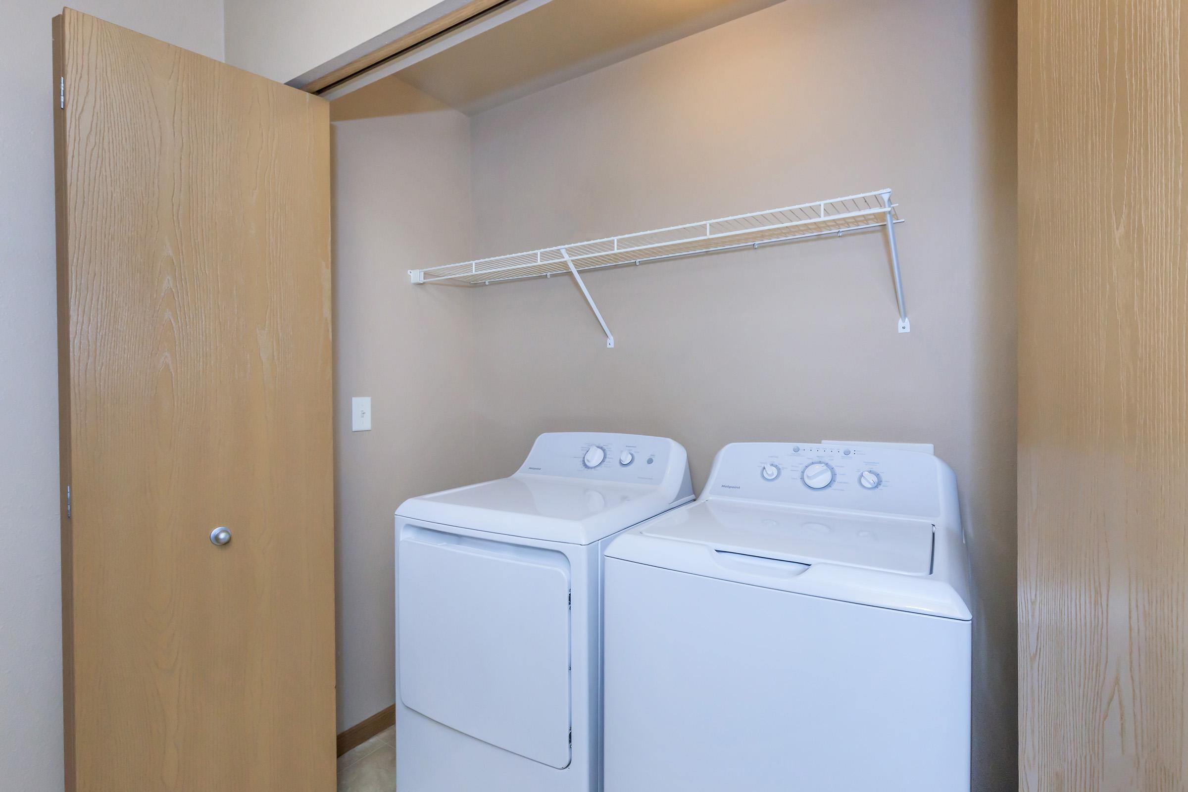 A laundry room featuring a stacked washer and dryer side by side, with a metal shelf above for storage. The room has light-colored walls and a wooden door that is partially open.