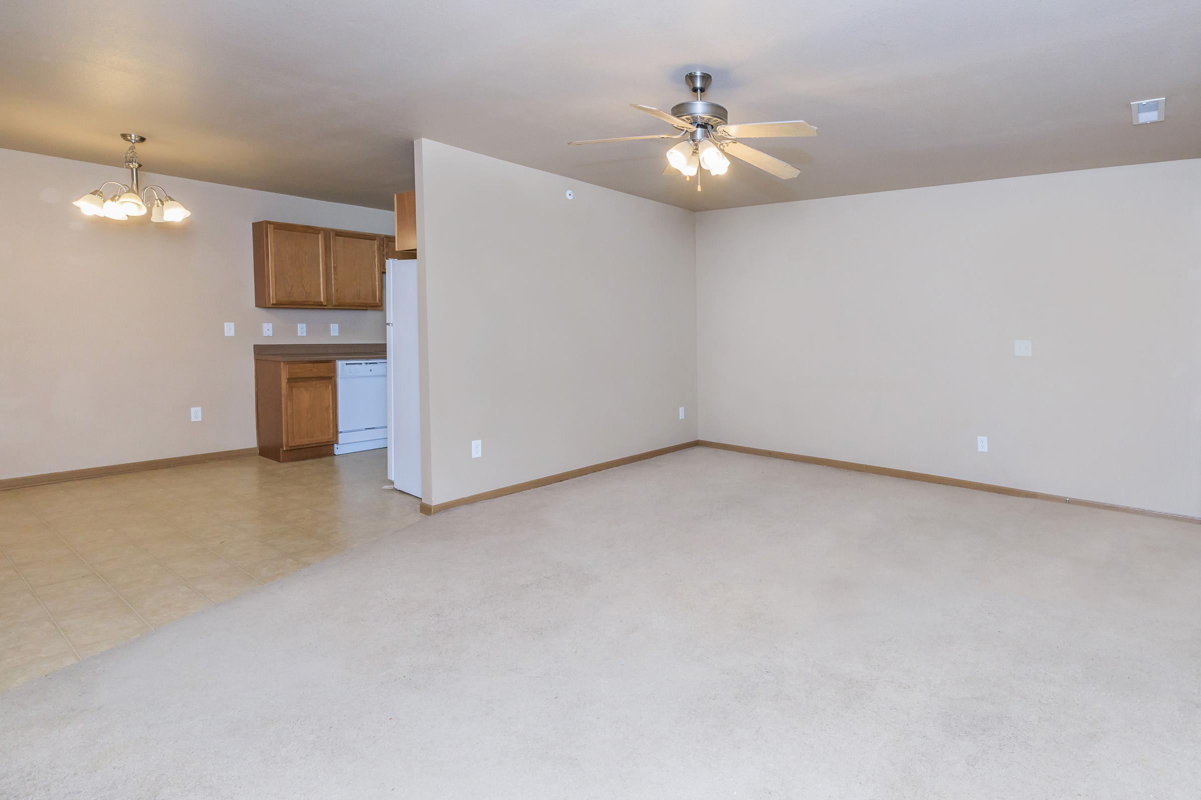 Spacious living area featuring beige walls, a ceiling fan, and light-colored flooring. An adjoining kitchen is visible with wooden cabinets and white appliances. The layout is open, creating a bright and airy ambiance suitable for various furniture arrangements.