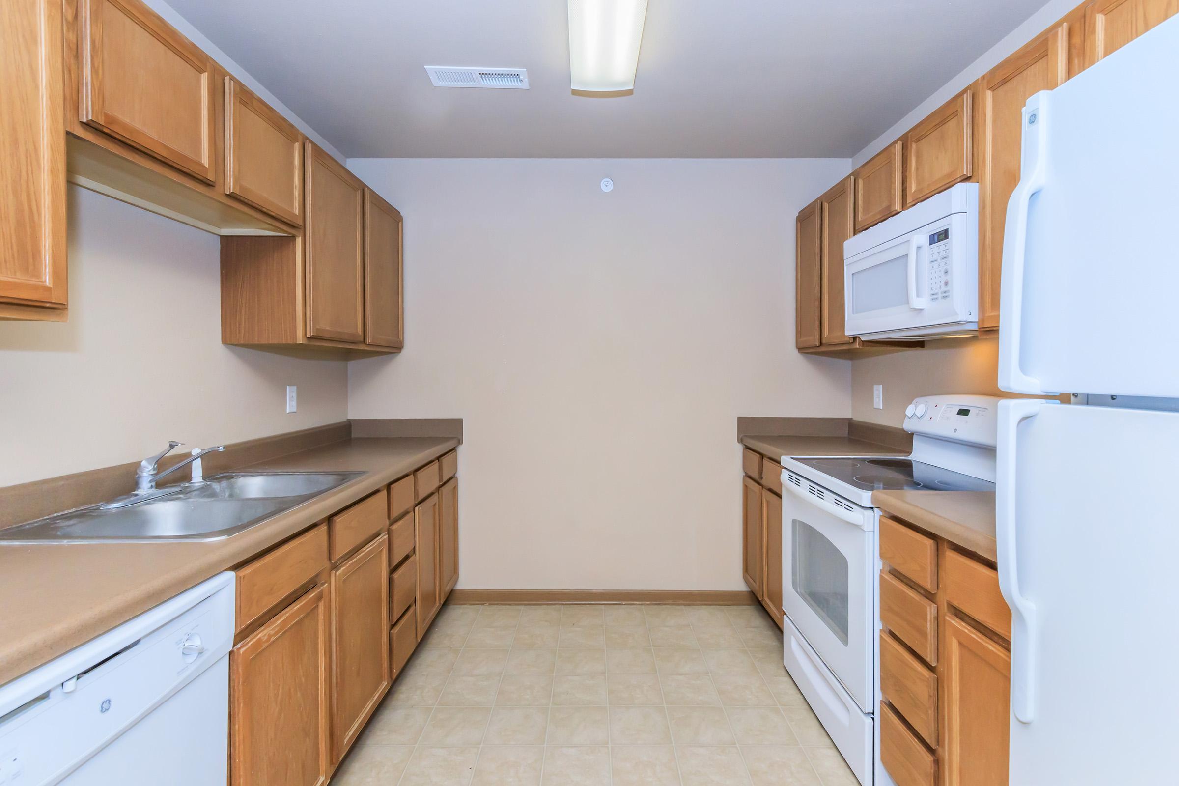 A modern kitchen featuring light brown wooden cabinets, beige walls, a double sink, and stainless steel appliances. The setup includes a white dishwasher, microwave, and oven, with a spacious layout and a neutral-colored floor. Natural light illuminates the room from a ceiling fixture.