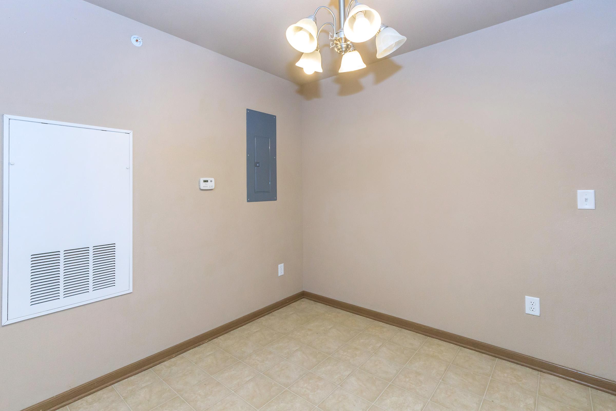 A plain, empty room with beige walls and tiled flooring. There is a ceiling light fixture with multiple bulbs, an electrical panel on one wall, a thermostat, and a vent in the corner. The room has no furniture or decorations, giving it a spacious and unoccupied appearance.