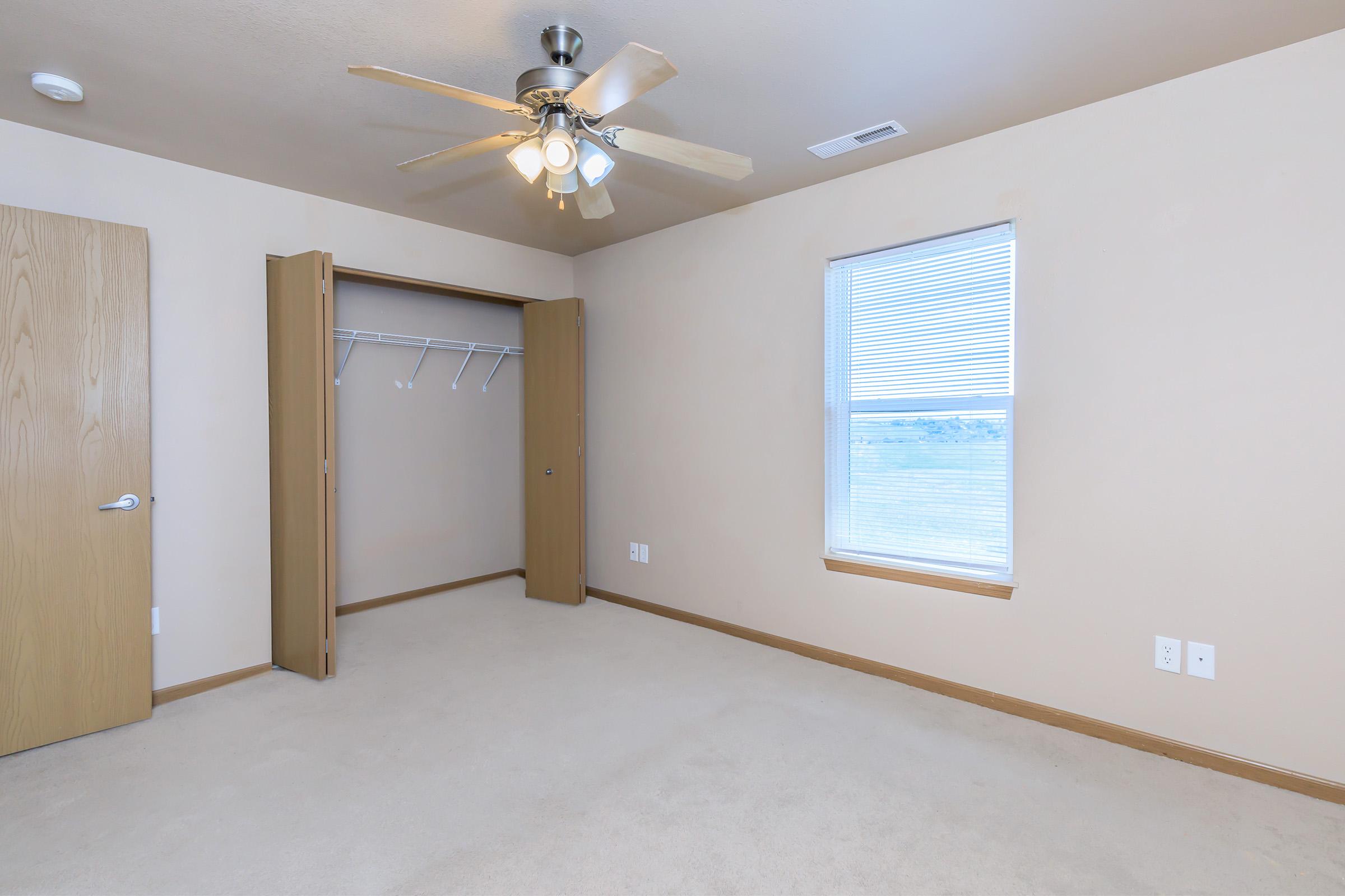 Empty room with light beige walls and carpet. A ceiling fan is mounted above, and there is a window with blinds that lets in natural light. An open closet with hanging rods is on the left side, and the room has no furniture, creating a spacious, minimalistic appearance.