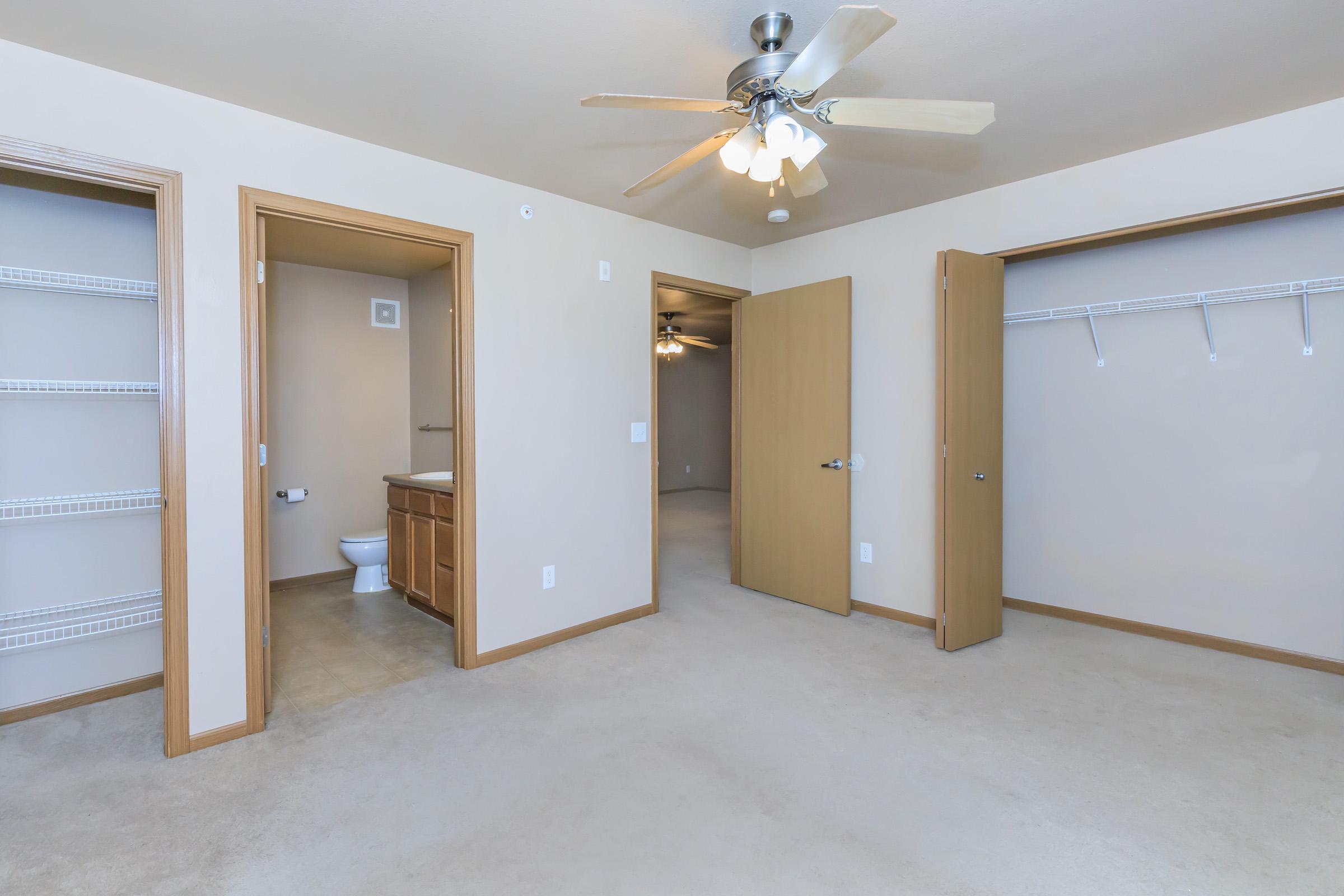 A spacious interior of an empty room featuring beige walls and carpet. Two open doors lead to a bathroom and a closet, while a ceiling fan hangs from the ceiling. There are shelves on one wall and ample natural light illuminating the space.