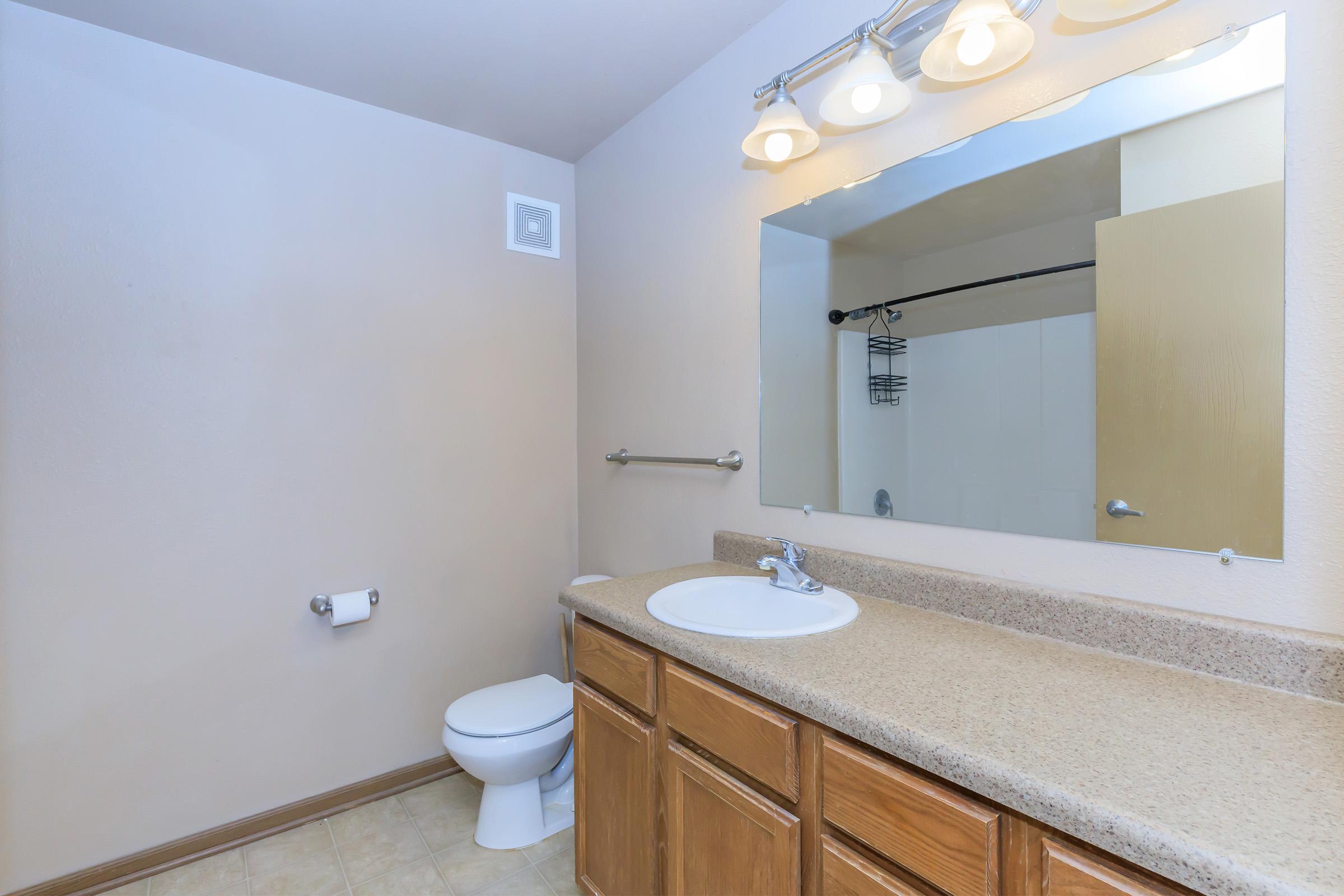 A clean and simple bathroom featuring a light-colored wall, a wooden vanity with a round sink, and a large mirror above it. There is a toilet beside the vanity, and the floor is tiled. Soft lighting from three ceiling fixtures illuminates the space.