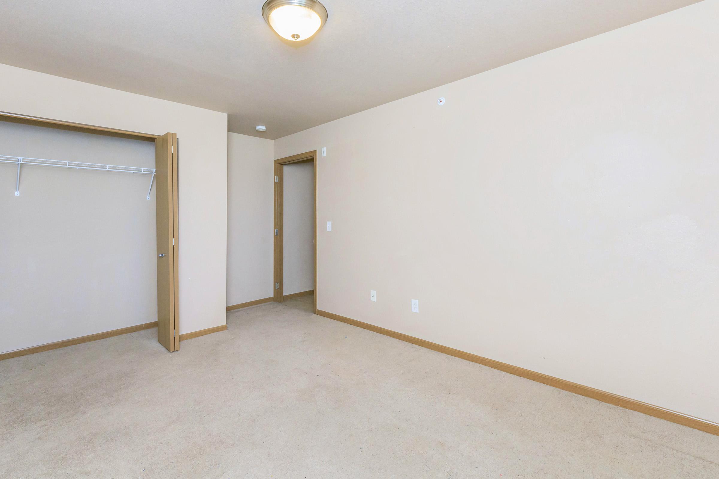 A spacious, empty room with beige walls and a light-colored carpet. There is a single closet with a sliding door on the left and a door leading to another room on the right. A ceiling light fixture is visible, providing soft illumination. The overall atmosphere is clean and minimalistic.