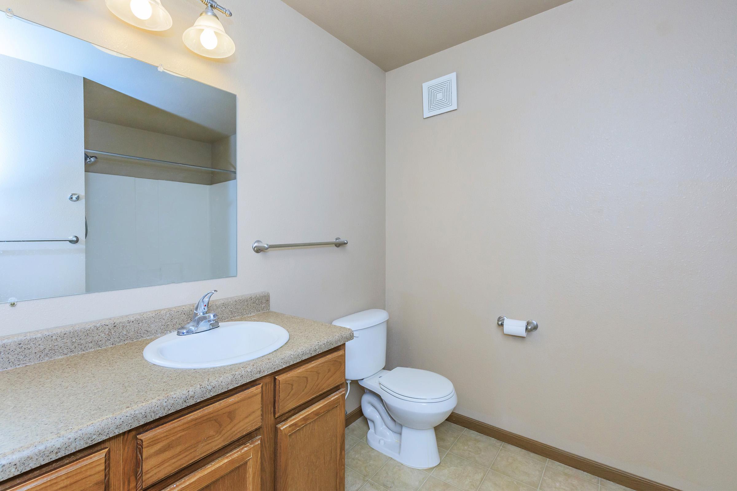 A bathroom featuring a light-colored wall, a mirror above a sink with a faucet, a toilet, and a towel bar. The countertop is beige, and the flooring has a light tile pattern. Soft lighting from two wall sconces illuminates the space, creating a clean and neutral atmosphere.