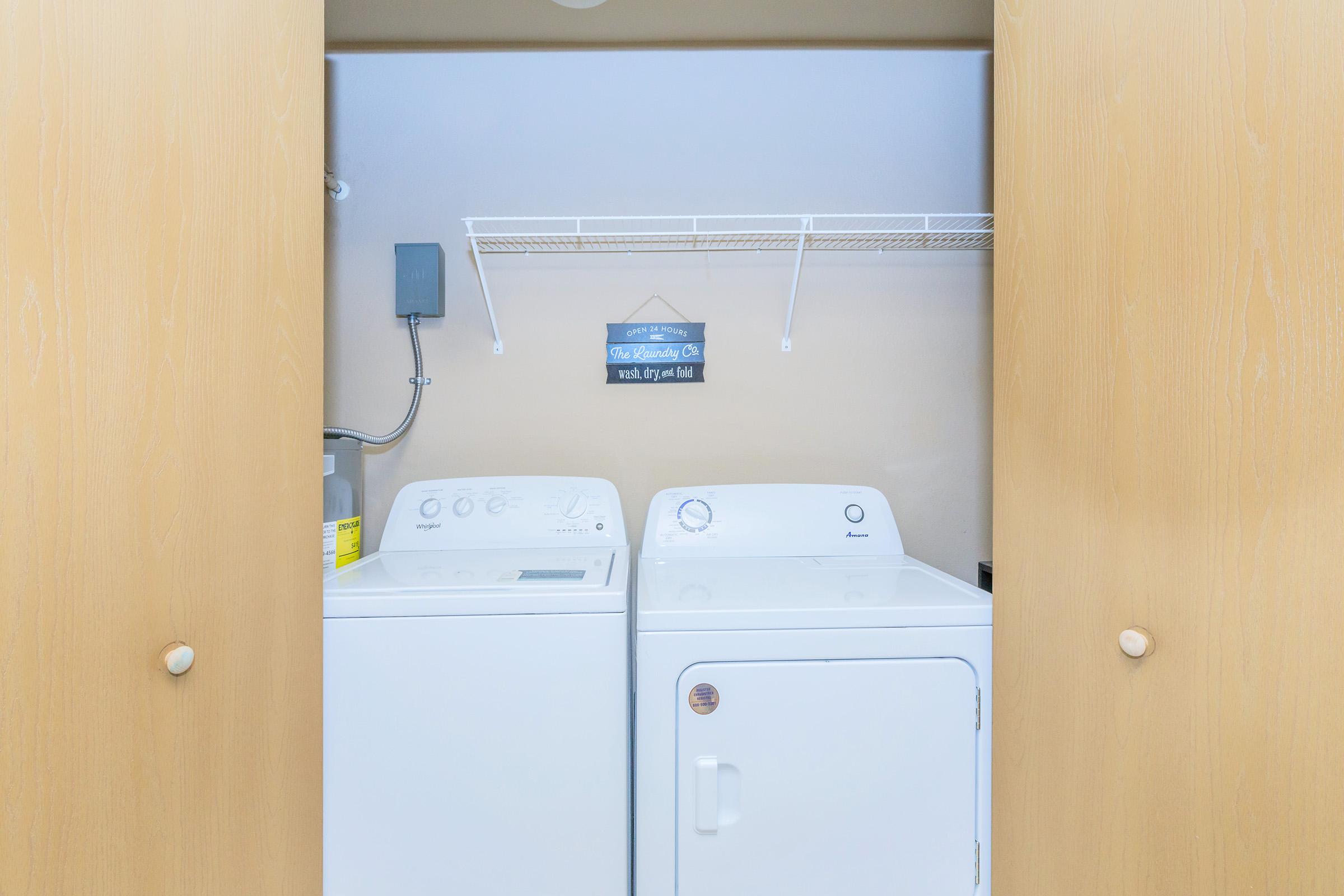 A clean laundry room featuring a washing machine and dryer side by side, enclosed by wooden cabinet doors. Above the appliances is a wire shelf with a decorative sign. The walls are painted in neutral tones, creating a tidy and organized space for laundry chores.