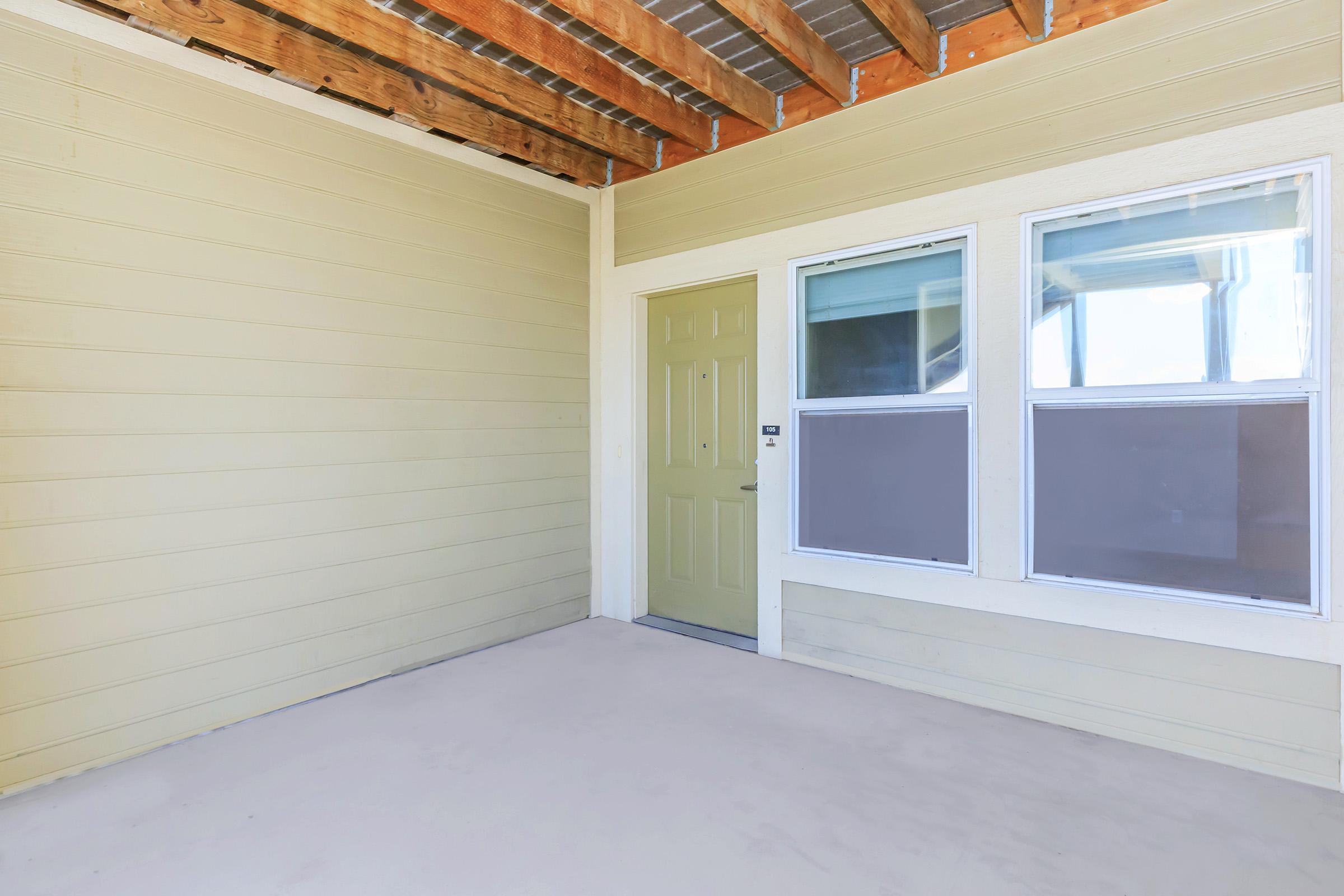 Empty porch area with a light yellow exterior wall and a green front door. Two windows with panes are visible, one partially covered. The ceiling has exposed wooden beams, and the floor is a plain light color. The space is well-lit and appears to be freshly painted.