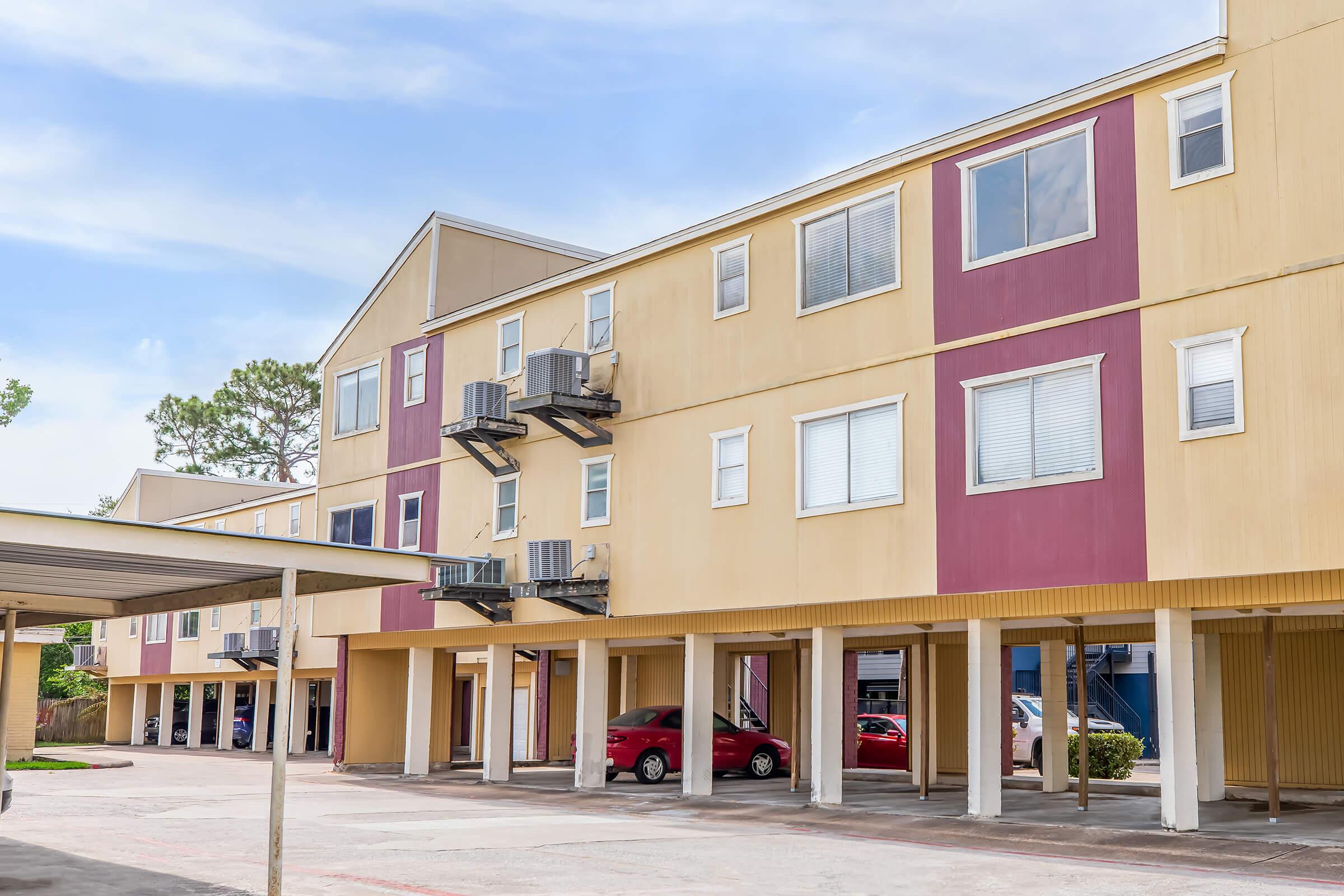 Multi-story building with beige and maroon exterior. Ground-level parking area is visible, featuring several parked cars. Air conditioning units are mounted on the walls. Clear blue sky is in the background, adding to the setting's bright and inviting atmosphere.