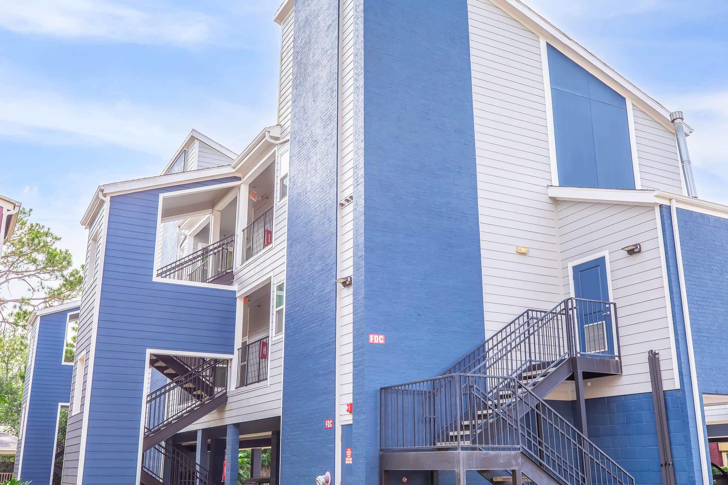 Exterior view of a modern multi-story blue apartment building with white siding. The building features a staircase leading to upper levels, balconies with red doors, and large windows. The sky is clear, adding to the bright and inviting atmosphere.