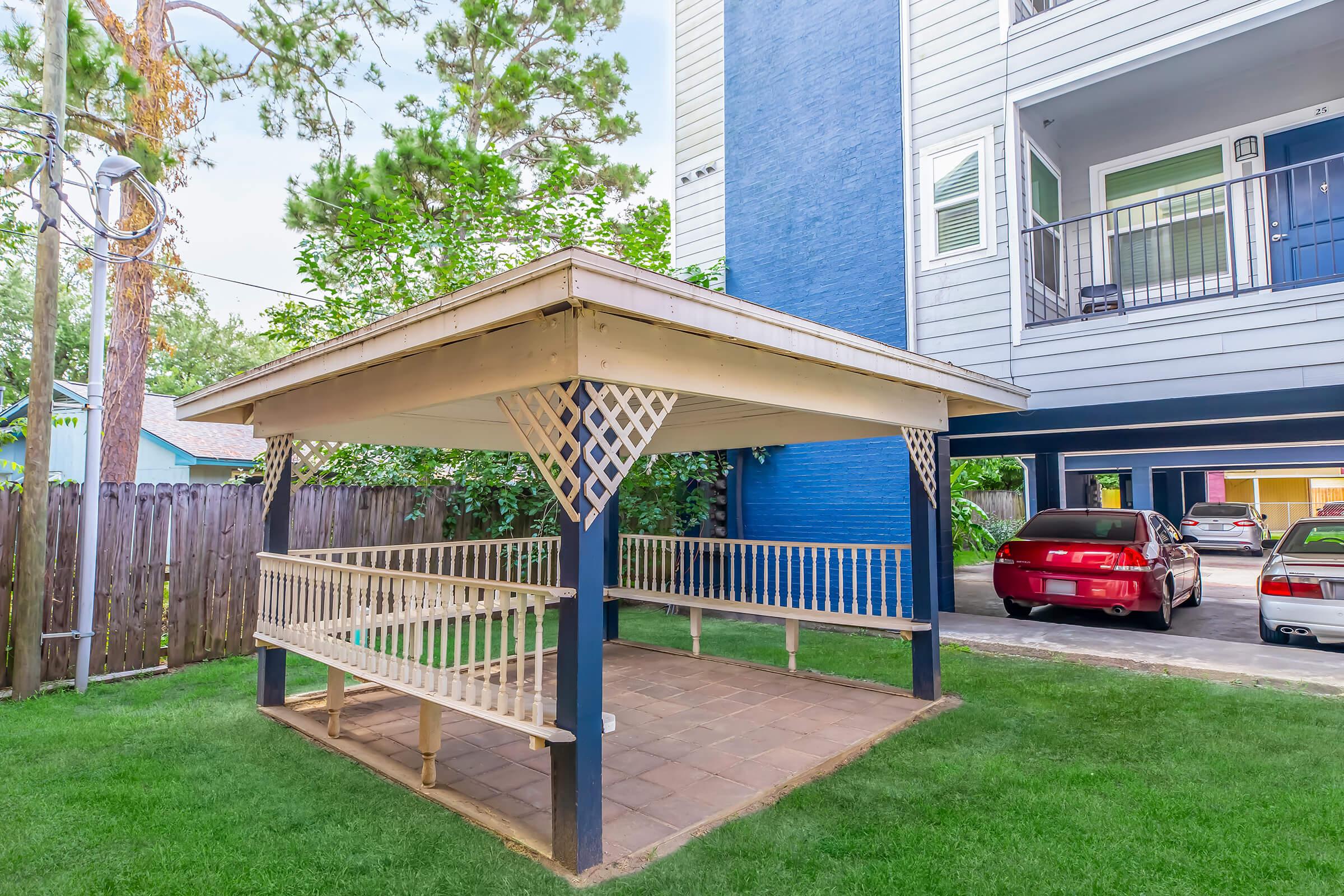 A gazebo with a wooden bench seating area, set in a grassy courtyard next to a multi-story building. The surrounding area features trees and a wooden fence. A couple of parked cars are visible nearby.