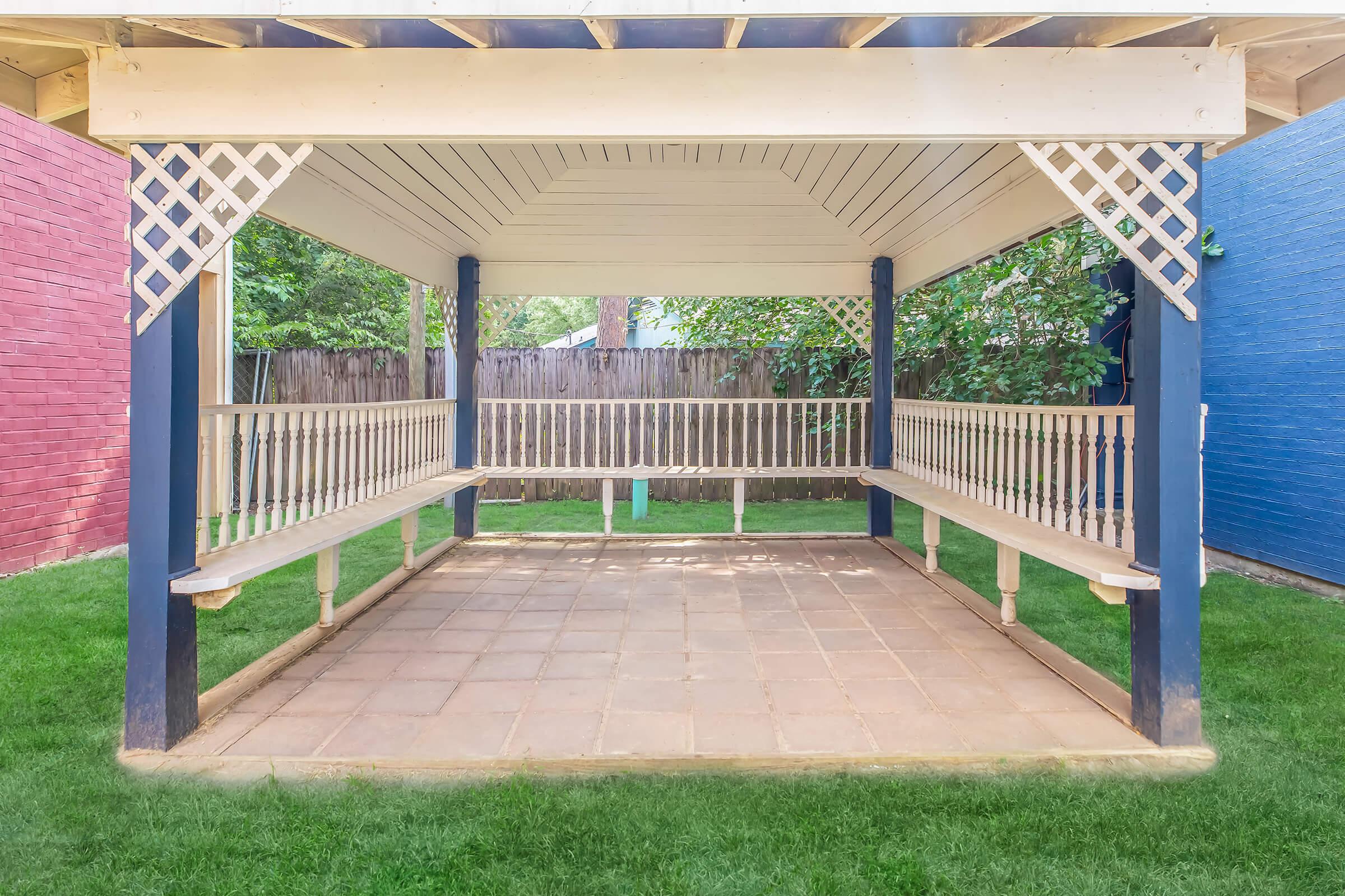 A spacious outdoor gazebo with benches on either side, set on a tiled floor. The gazebo features a sloped roof and decorative lattice accents. Surrounded by green grass and trees, it provides a shaded area for relaxation or gatherings. Two colorful walls in the background add charm to the setting.