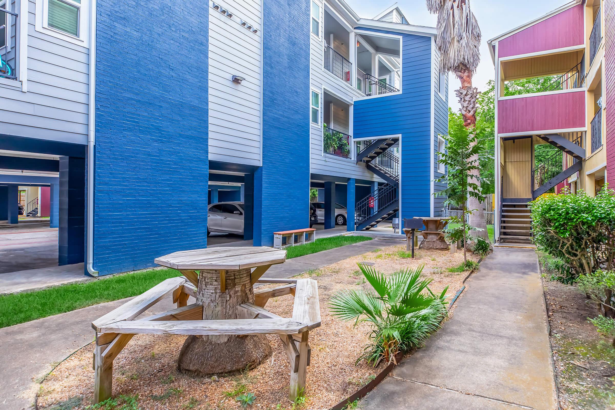 A colorful apartment complex featuring two multi-level buildings with blue and red exteriors. In the foreground, there is a wooden picnic table made from a tree stump, surrounded by a landscaped area with palm plants and a paved walkway. Parking is visible in the background under the buildings.
