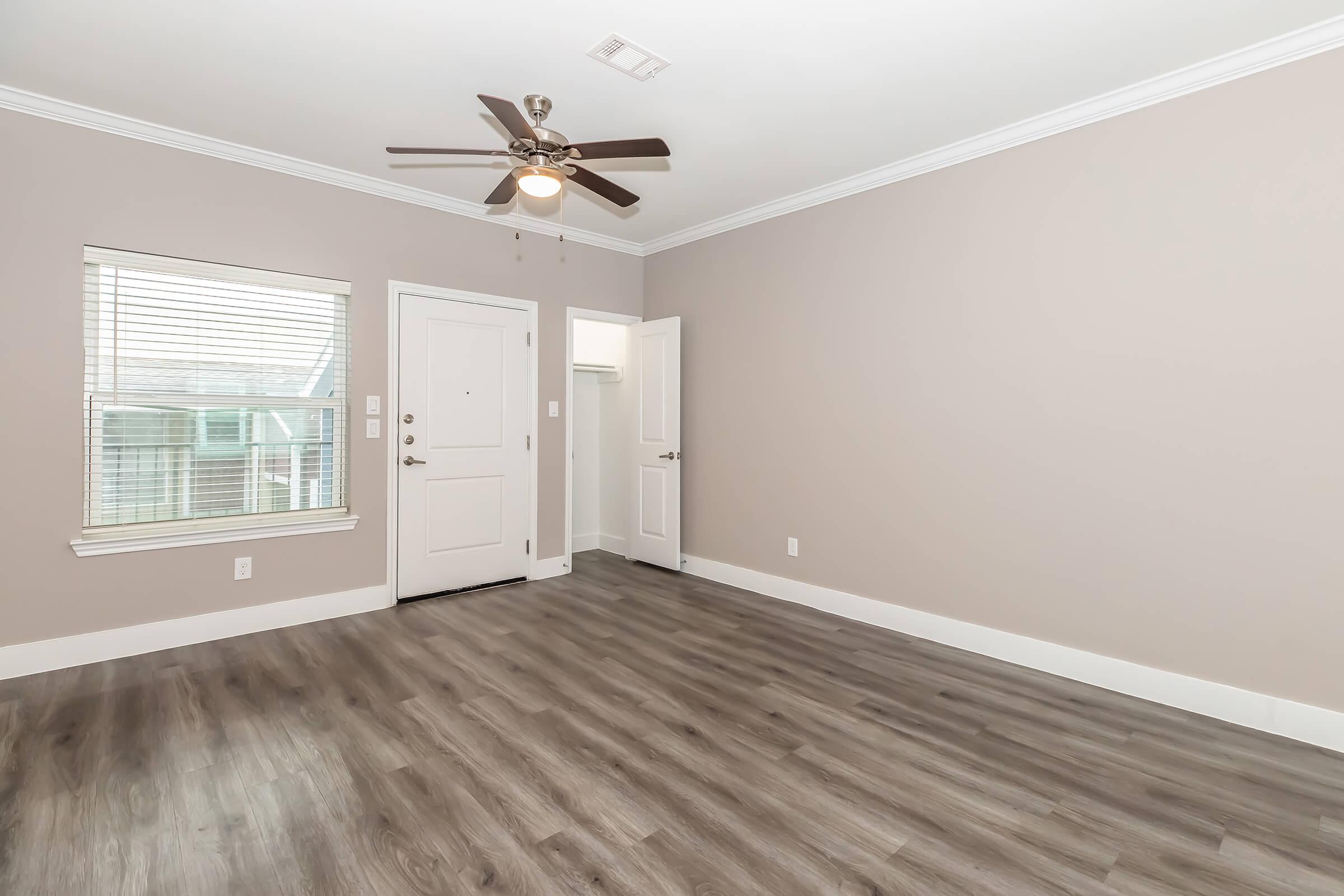 Empty room with light brown walls and a wooden floor. A ceiling fan is overhead, and there's a window allowing natural light. A door leads to another area, and another door is slightly ajar, indicating access to a closet. The space is clean and uncluttered, perfect for personalized decoration.