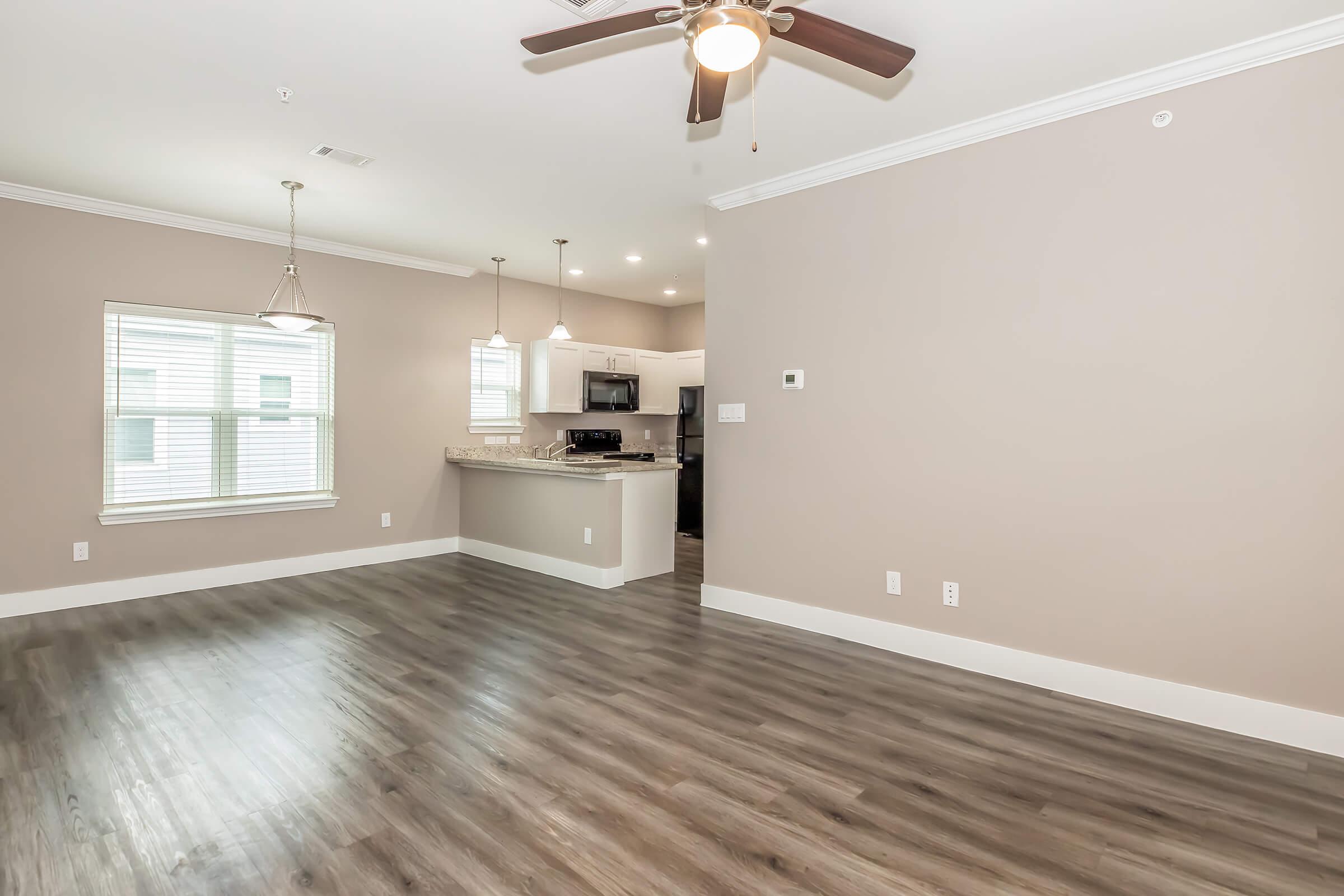 A spacious and modern living area with light brown walls and hardwood flooring. It features a ceiling fan, a large window providing natural light, and an open kitchen area visible in the background, equipped with dark cabinets and appliances. The overall ambiance is bright and inviting.