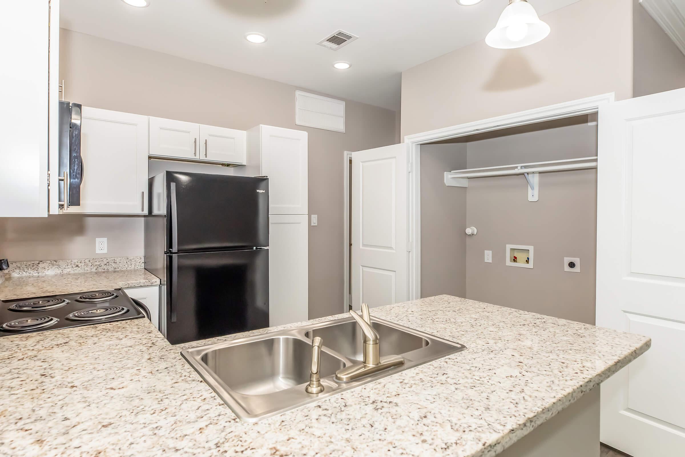 A modern kitchen featuring a granite countertop with a double sink, a black refrigerator, and white cabinets. The stove is set into the counter. There is a small closet space with a shelf and hanging rod visible in the background. The walls are painted in a neutral color, enhancing the bright, airy feel of the space.