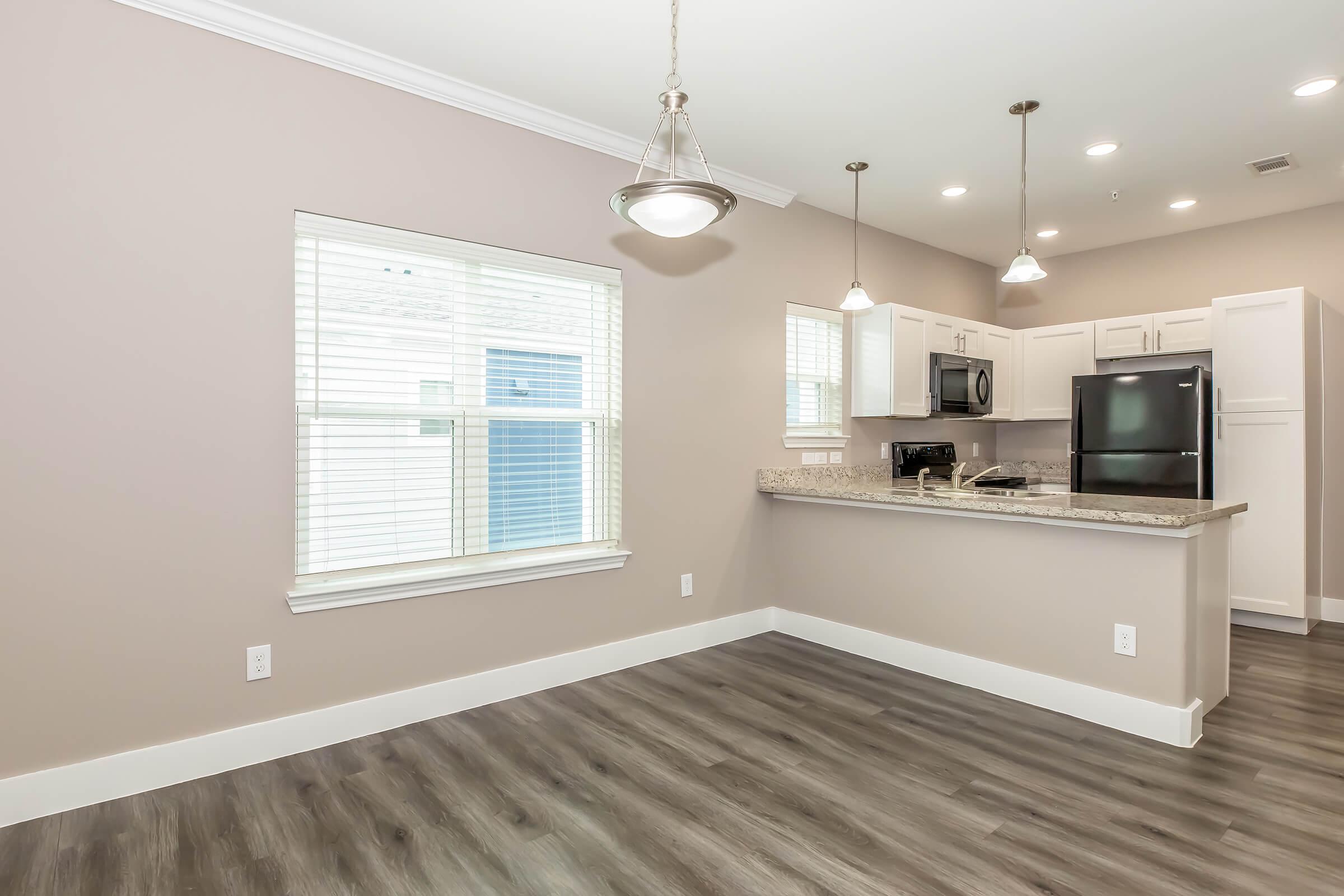 A modern kitchen and living area featuring light-colored walls, a window with blinds, and a view of the kitchen area with stainless steel appliances, granite countertops, and pendant lighting. The flooring is a dark wood laminate, creating a warm and inviting atmosphere.