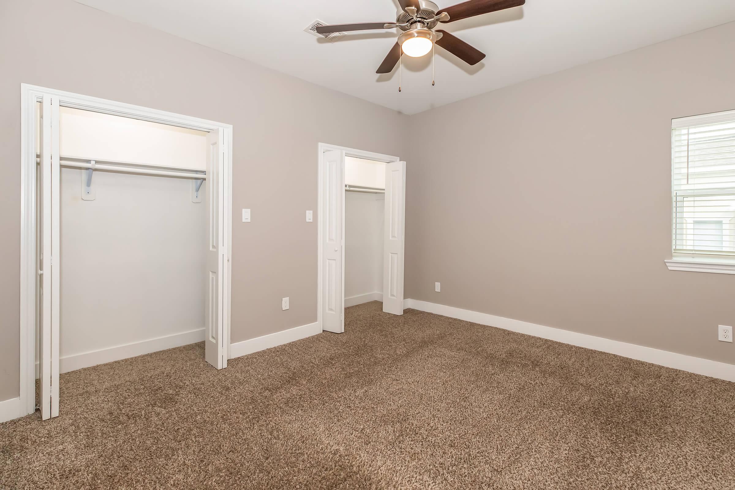 A spacious, empty bedroom featuring light beige walls and plush carpeting. Two open closet doors are visible on one wall, and a ceiling fan with wooden blades hangs above. A window allows natural light to enter, enhancing the bright and airy feel of the room.