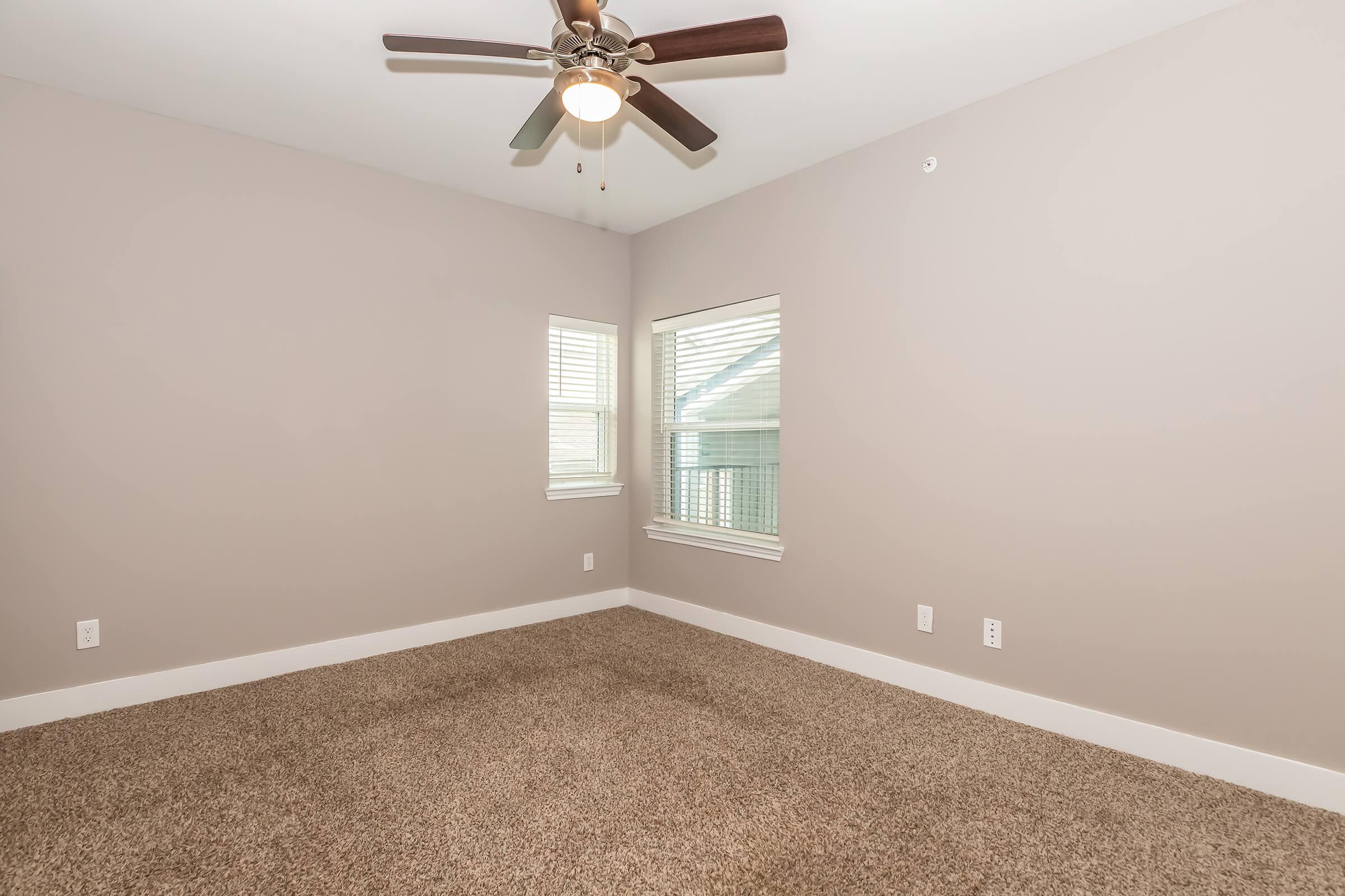 A spacious and unfurnished room with light beige walls and plush carpet. It features two windows with blinds allowing natural light, a ceiling fan with wooden blades, and white baseboards. The overall atmosphere is bright and inviting, ideal for personal decoration.