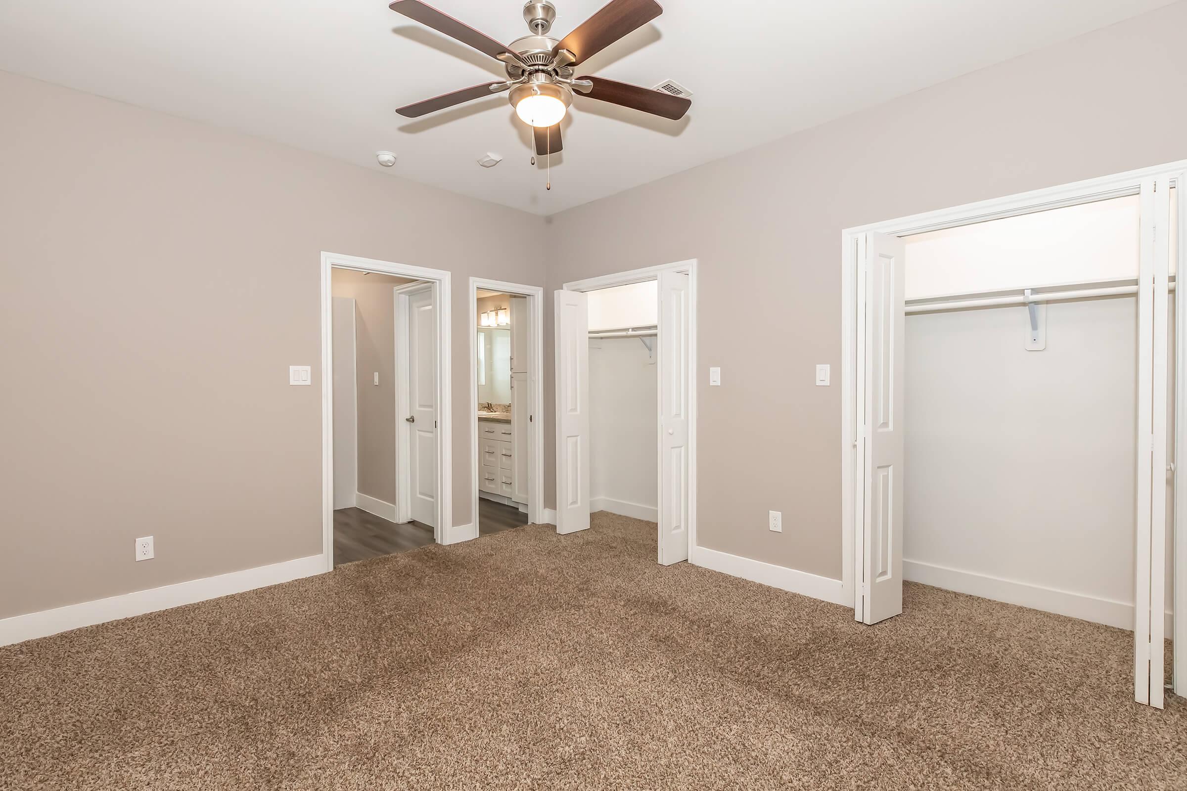 A spacious room featuring a ceiling fan, light gray walls, and plush carpet. There are two open doorways leading to closets on either side, and a doorway at the back appears to lead to a bathroom area. The overall design is modern and inviting.
