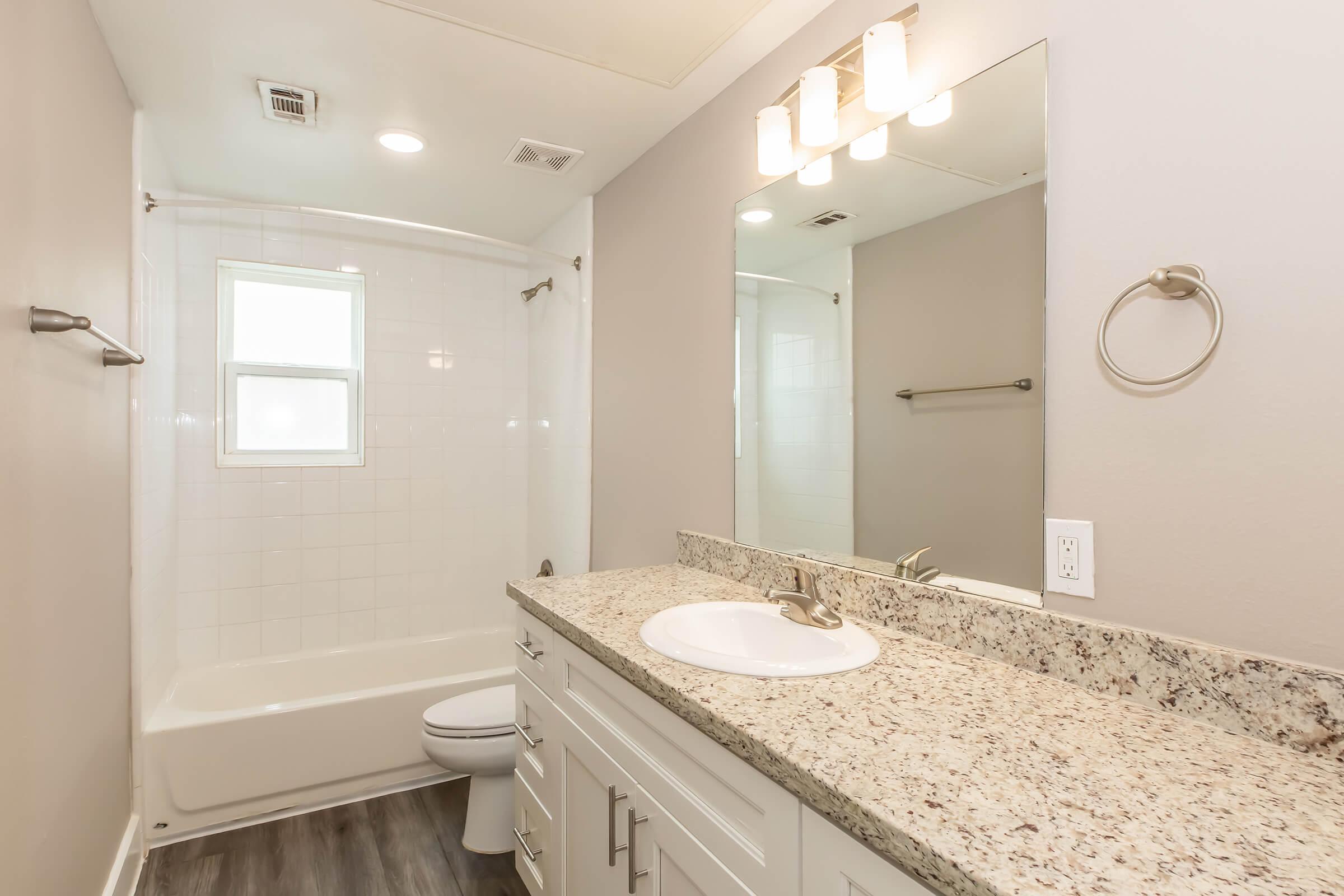 A modern bathroom featuring a white bathtub and shower combination, a granite countertop with a sink, a large mirror with three light fixtures above, and gray walls. The room includes a toilet and a towel ring, with natural light coming from a small window.