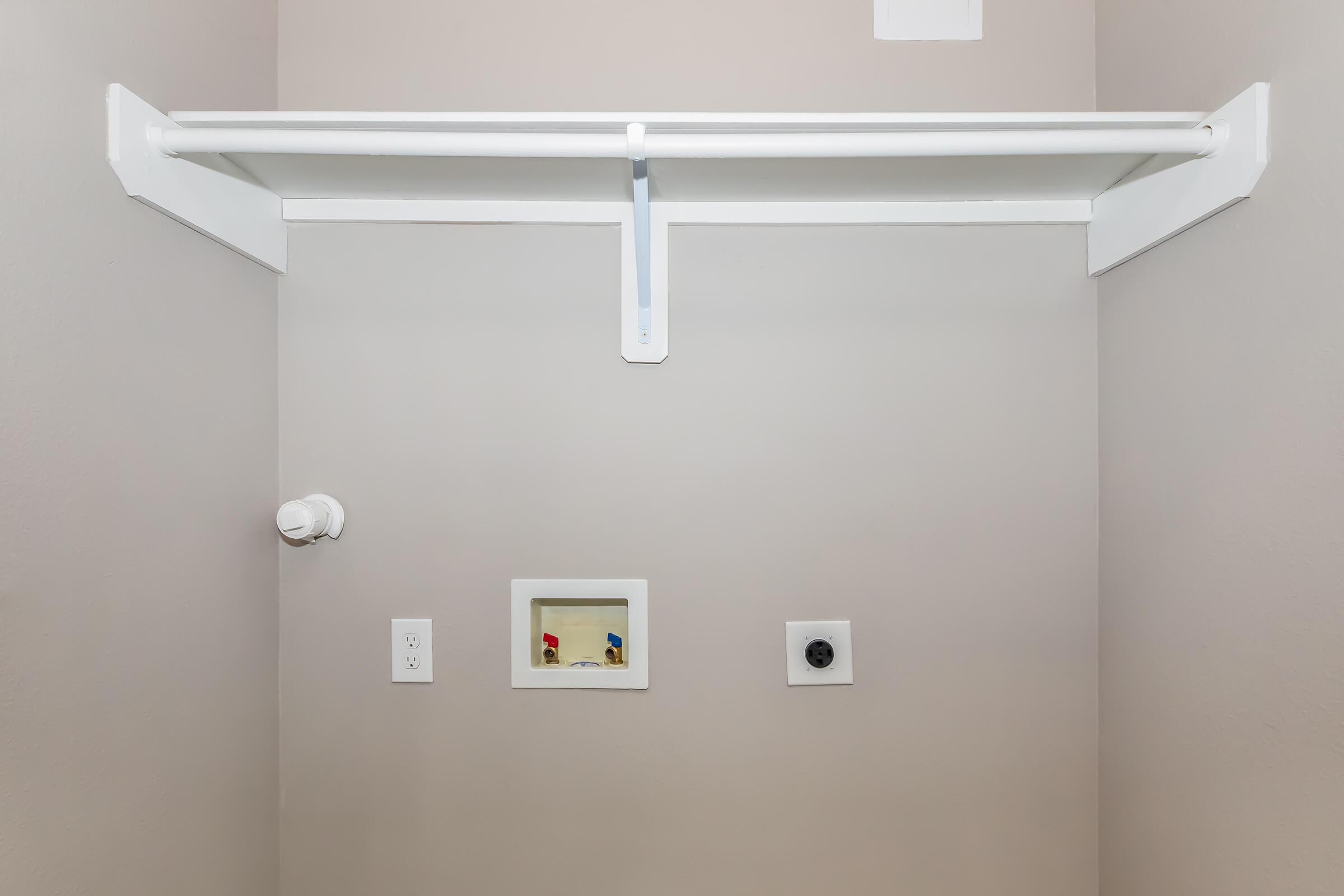 A clean, empty closet with a white shelf mounted on the wall. Below the shelf, there are two small wall fixtures: one is a power outlet, and the other is a small opening with visible wiring. The walls are painted a neutral color, creating a simple and minimalist appearance.