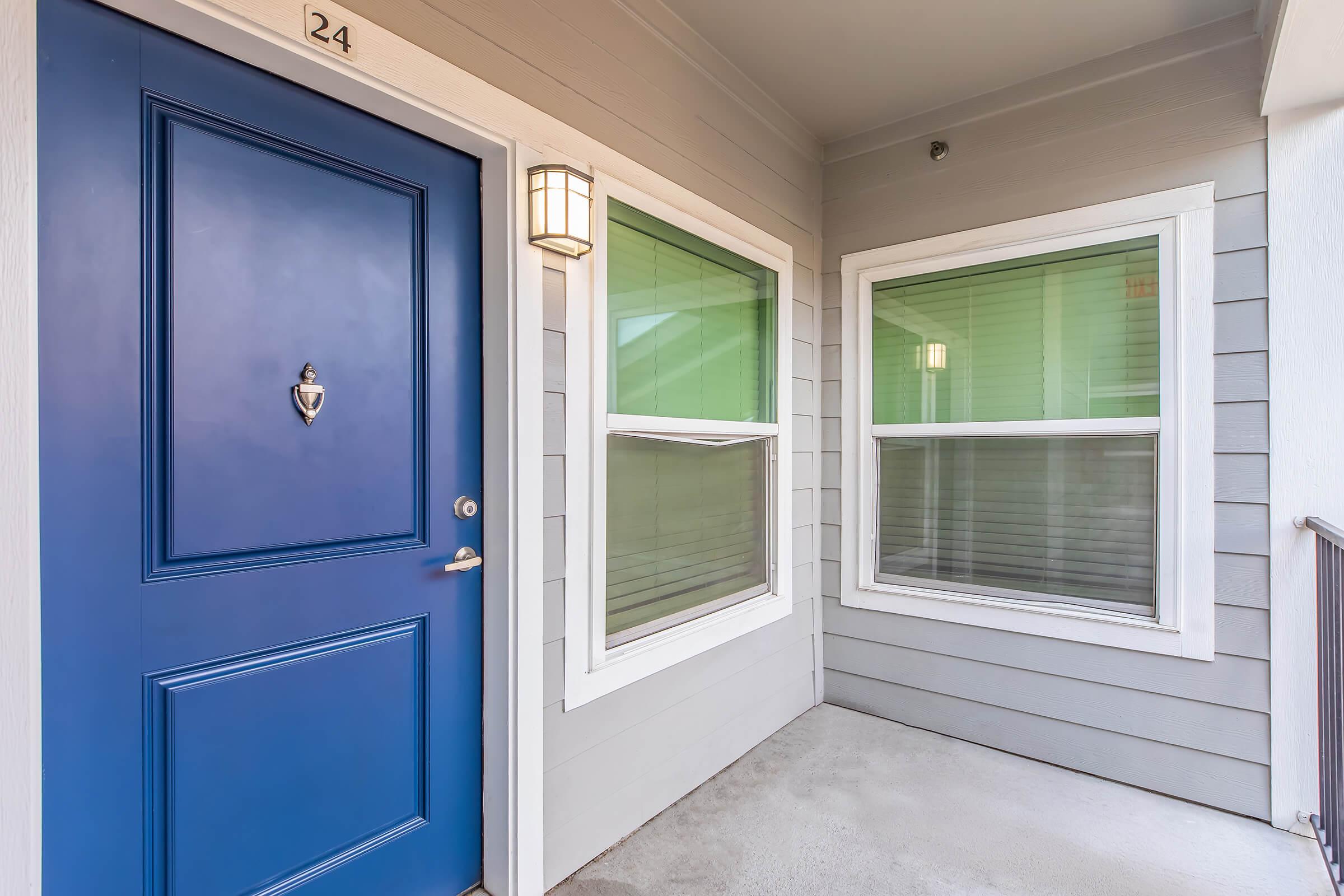 A blue front door with a silver doorknob and a doorbell, located next to two windows with green trims. The exterior features light gray siding and a small porch area, providing a welcoming entrance to the home.