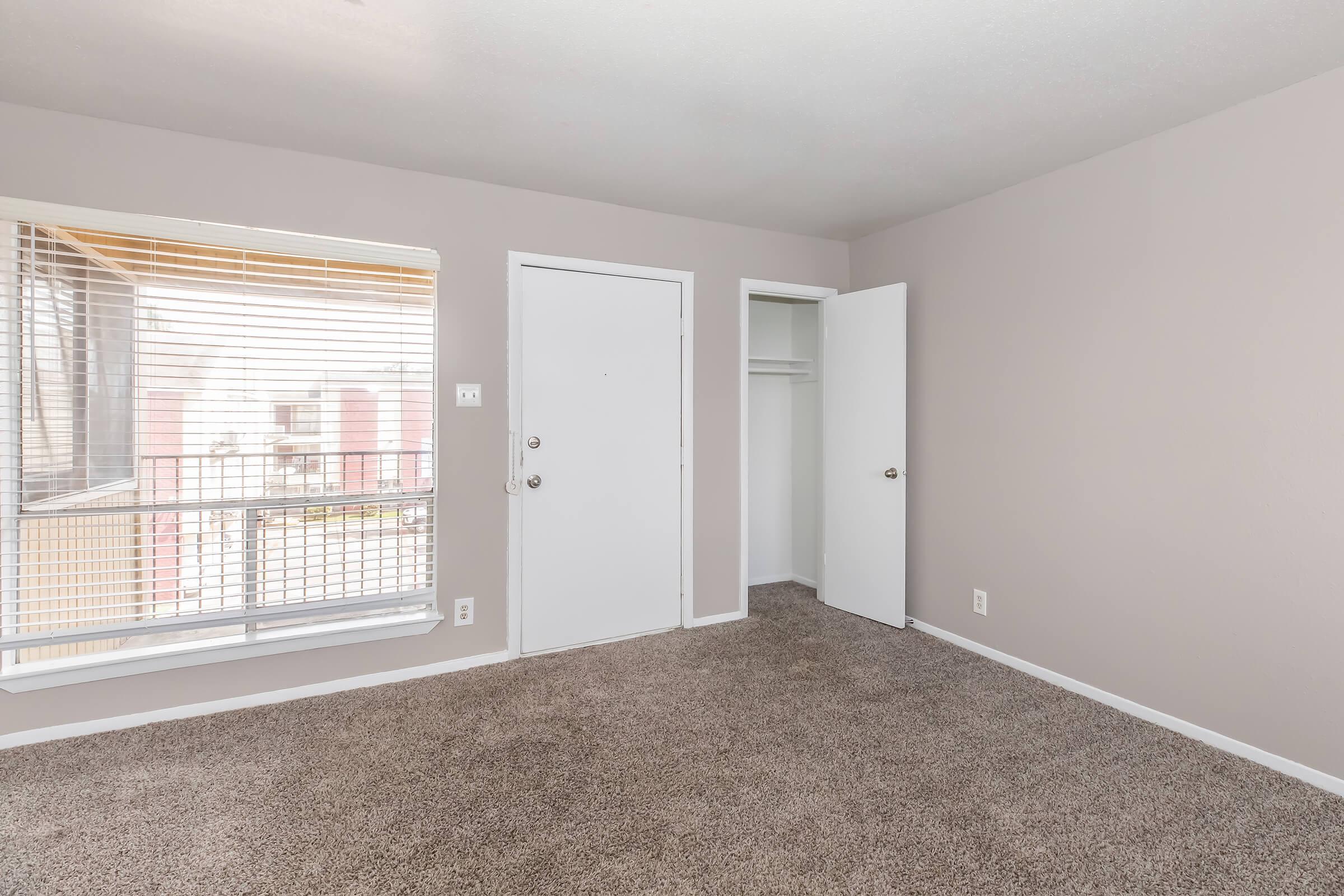 Empty room with light beige walls and carpet, featuring a large window with blinds, a white door leading outside, and a closet door. The room is well-lit and spacious, highlighting the neutral decor and clean lines.