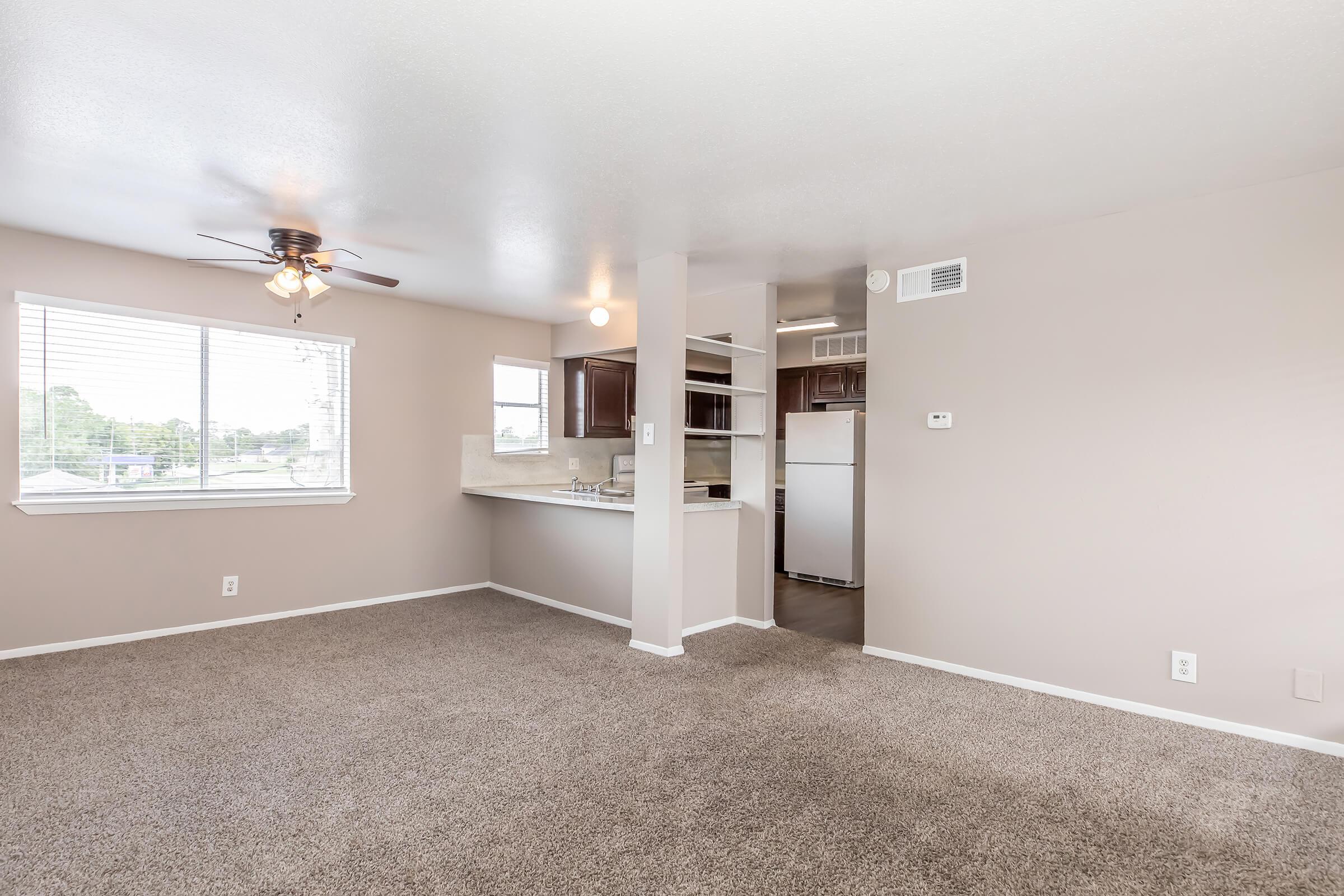 Spacious living area with beige walls, light carpet, and a ceiling fan. A large window allows natural light, showing part of a kitchen with dark wood cabinetry and white appliances in the background. The layout is open, creating a bright and inviting atmosphere.